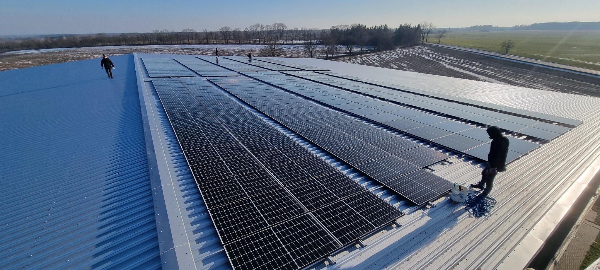 Rząd paneli słonecznych na blaszanym dachu budynku gospodarczego. Widoczni monterzy podczas pracy. W tle zaśnieżone pola i drzewa.