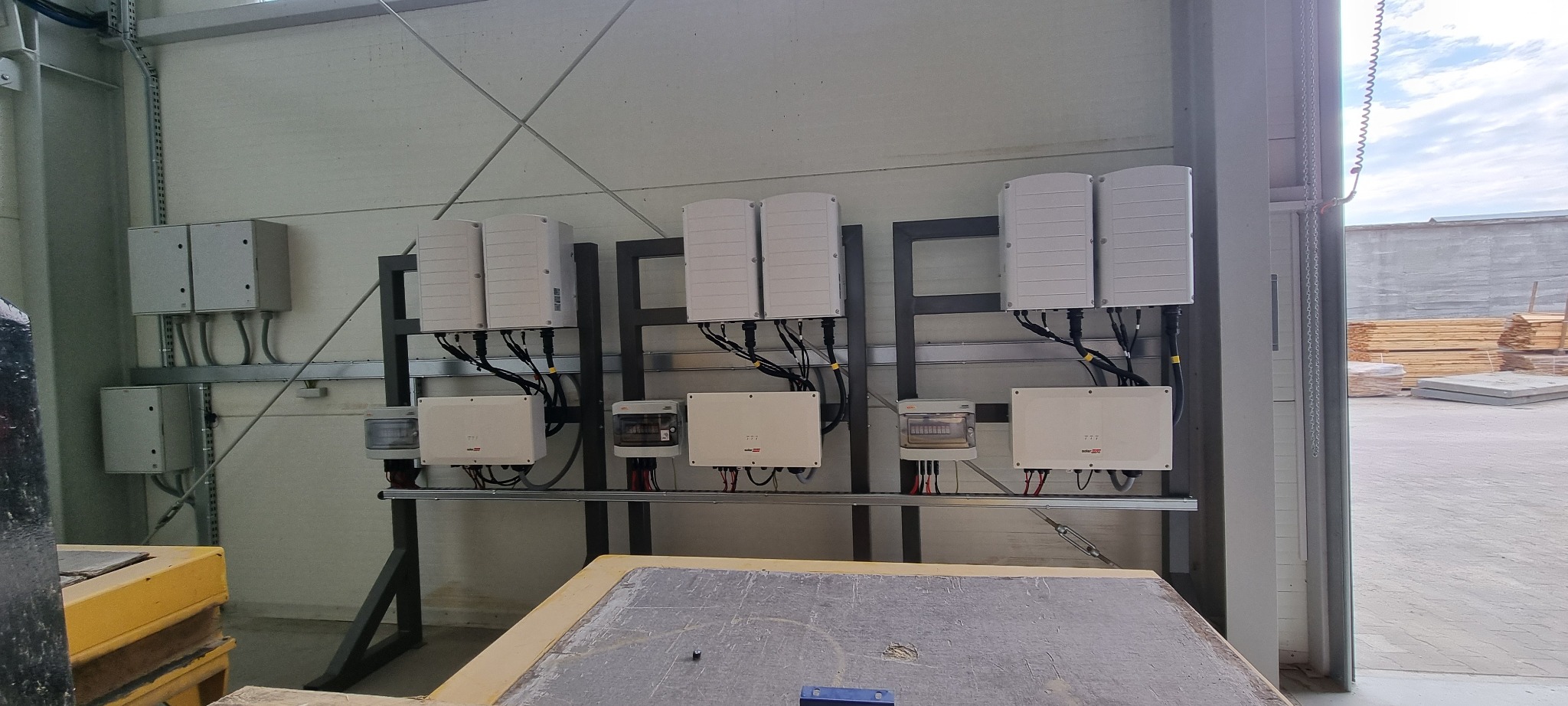 Rząd białych inwerterów i liczników na metalowej konstrukcji w hali, z widocznymi kablami i przewodami. W tle brama i składowane materiały budowlane.
