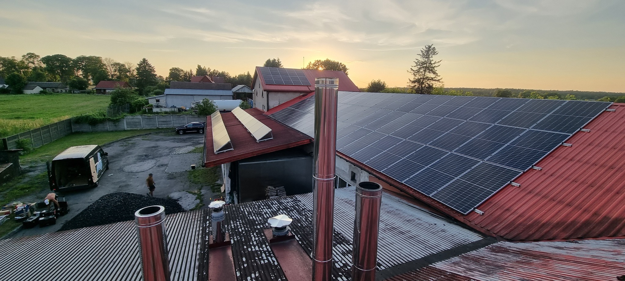 Instalacja paneli słonecznych na dachu budynku z blachy falistej, widok z góry, w tle krajobraz z drzewami i zachodzące słońce.
