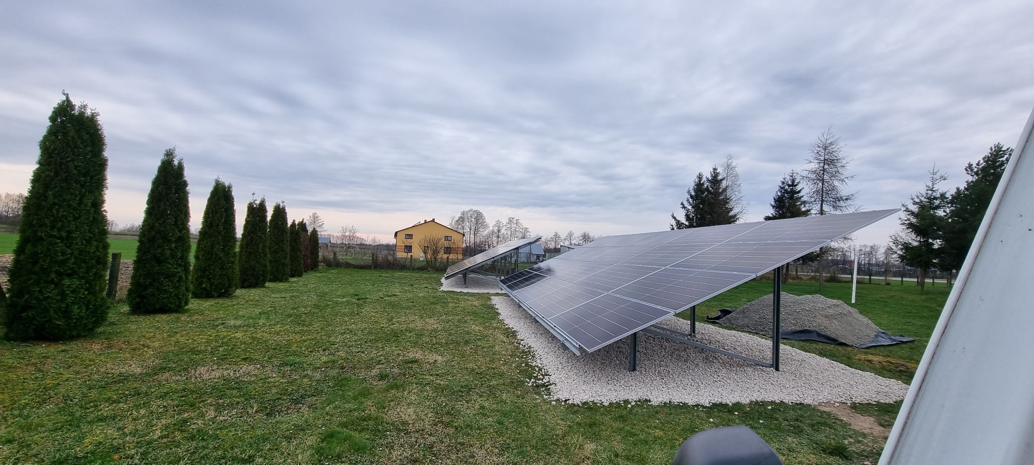 Dwie instalacje fotowoltaiczne na żwirowym podłożu, widok z boku, w tle dom i rząd tuj, pochmurne niebo. Część kadru zasłania biały element.