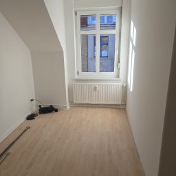 Sebastian Łachowski - Jasne, puste poddasze z oknem i drewnianą podłogą. Świeżo pomalowane ściany, grzejnik pod oknem. Kabel zasilający i rura leżą na podłodze. Widok na budynek zza okna.