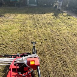 Sebastian Łachowski - Wertykulacja trawnika z użyciem wertykulatora elektrycznego. Widoczny pracownik z grabiami i zielona toaleta przenośna w tle. Trawa po zabiegu.