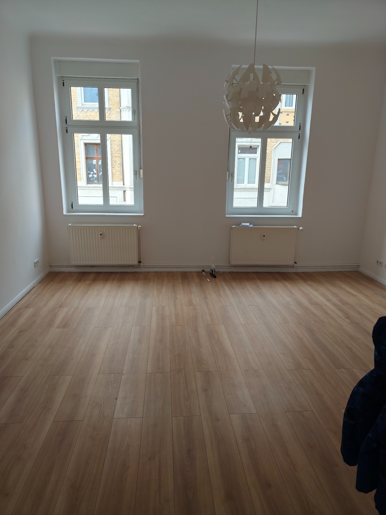 Jasne panele podłogowe w pustym pokoju z dwoma oknami i białymi ścianami. Wisząca lampa w stylu skandynawskim. Grzejniki pod oknami. Widok na budynki z zewnątrz.
