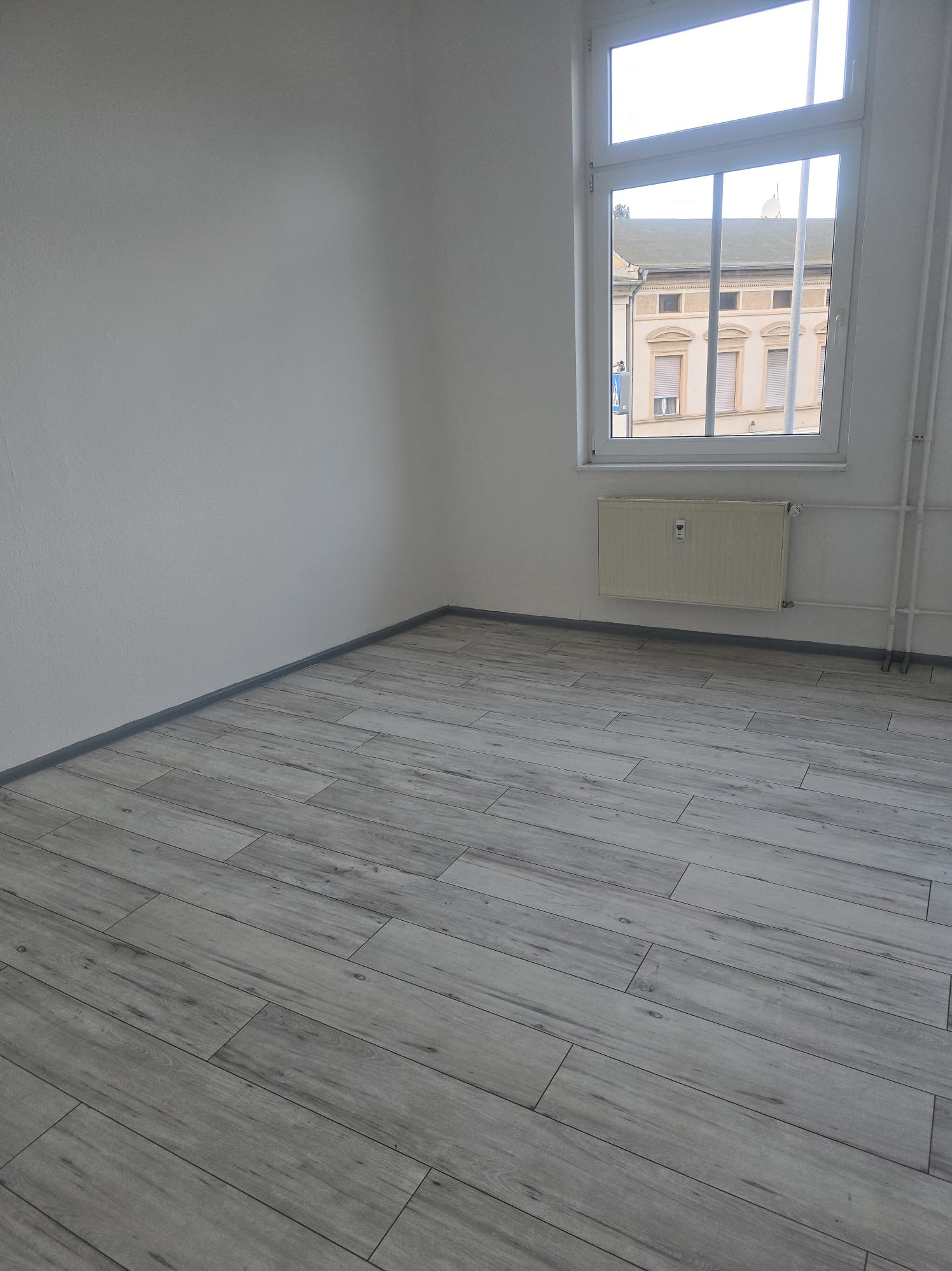 Jasne, puste wnętrze po remoncie z panelami podłogowymi imitującymi drewno, grzejnikiem pod oknem i świeżo pomalowanymi ścianami.