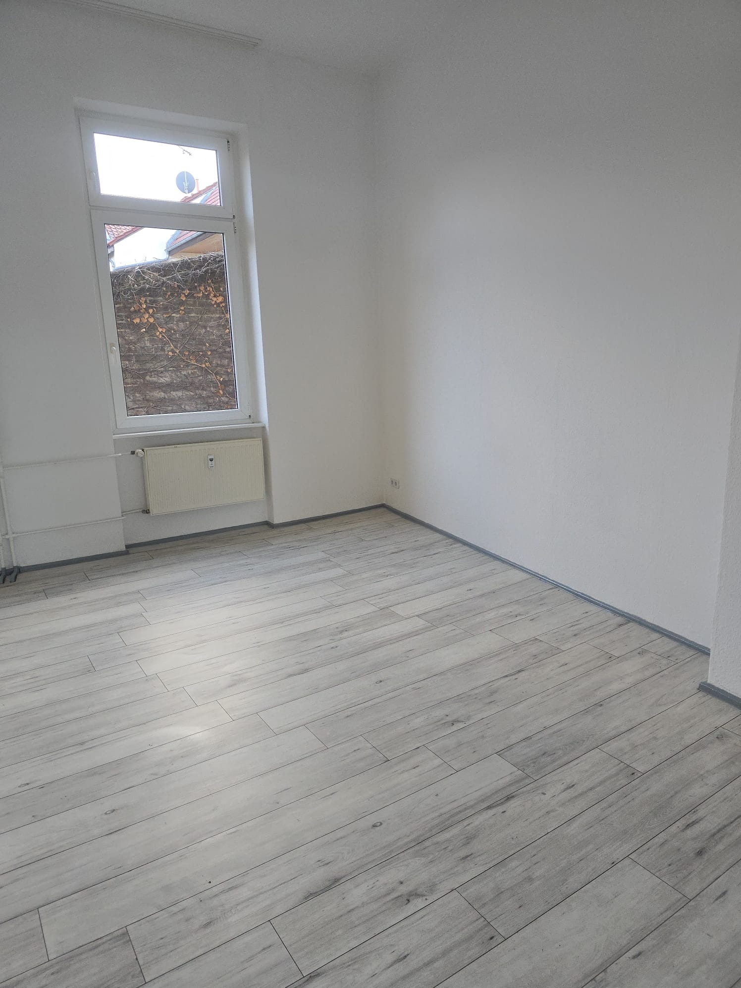 Jasne panele podłogowe w pustym, świeżo wyremontowanym pokoju z białymi ścianami i oknem. Widoczny grzejnik pod oknem. Minimalistyczny wystrój wnętrza.