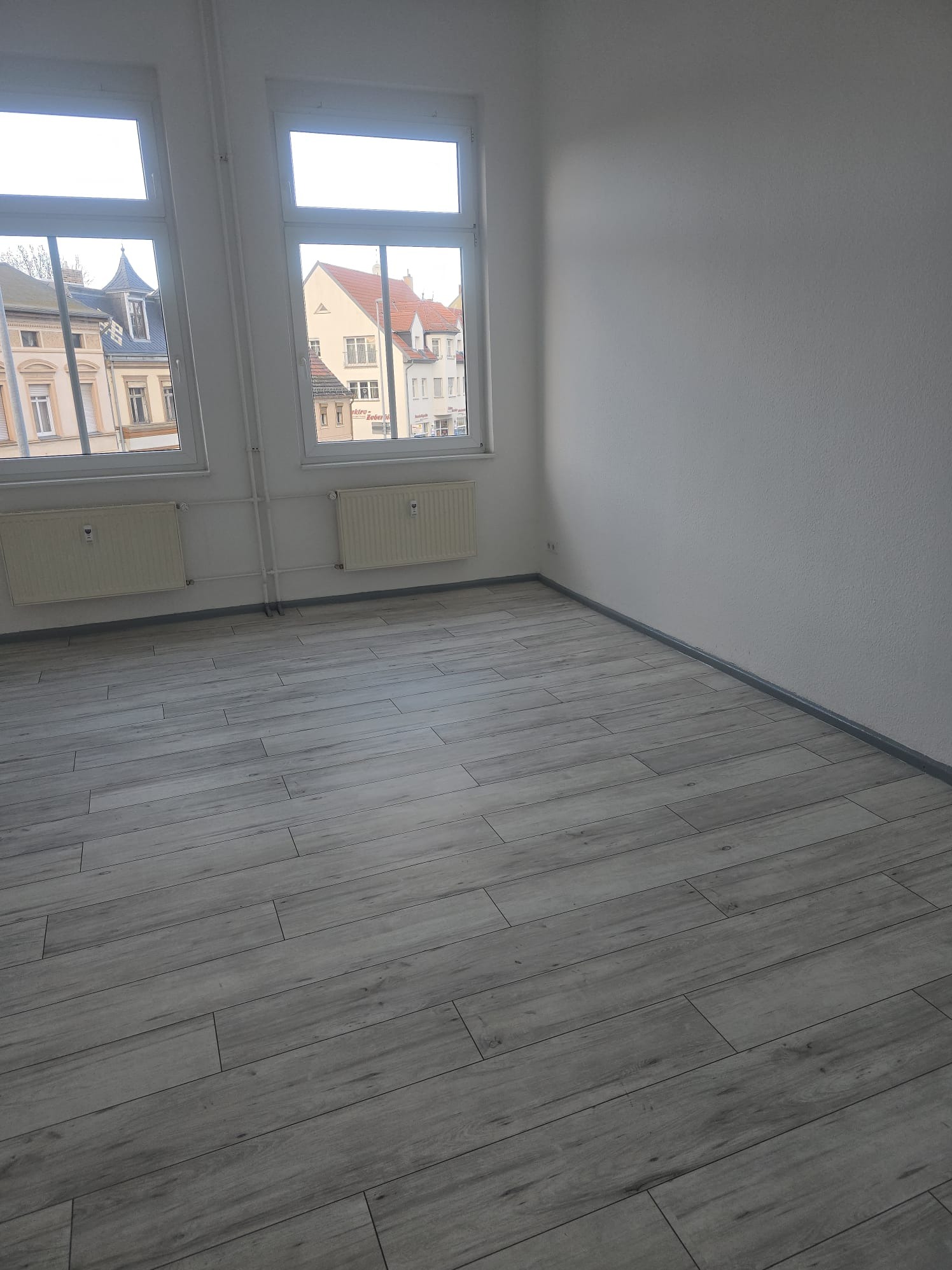 Jasne, wykończone wnętrze z panelami podłogowymi imitującymi drewno, białymi ścianami i dwoma oknami z widokiem na budynki. Grzejniki pod oknami.