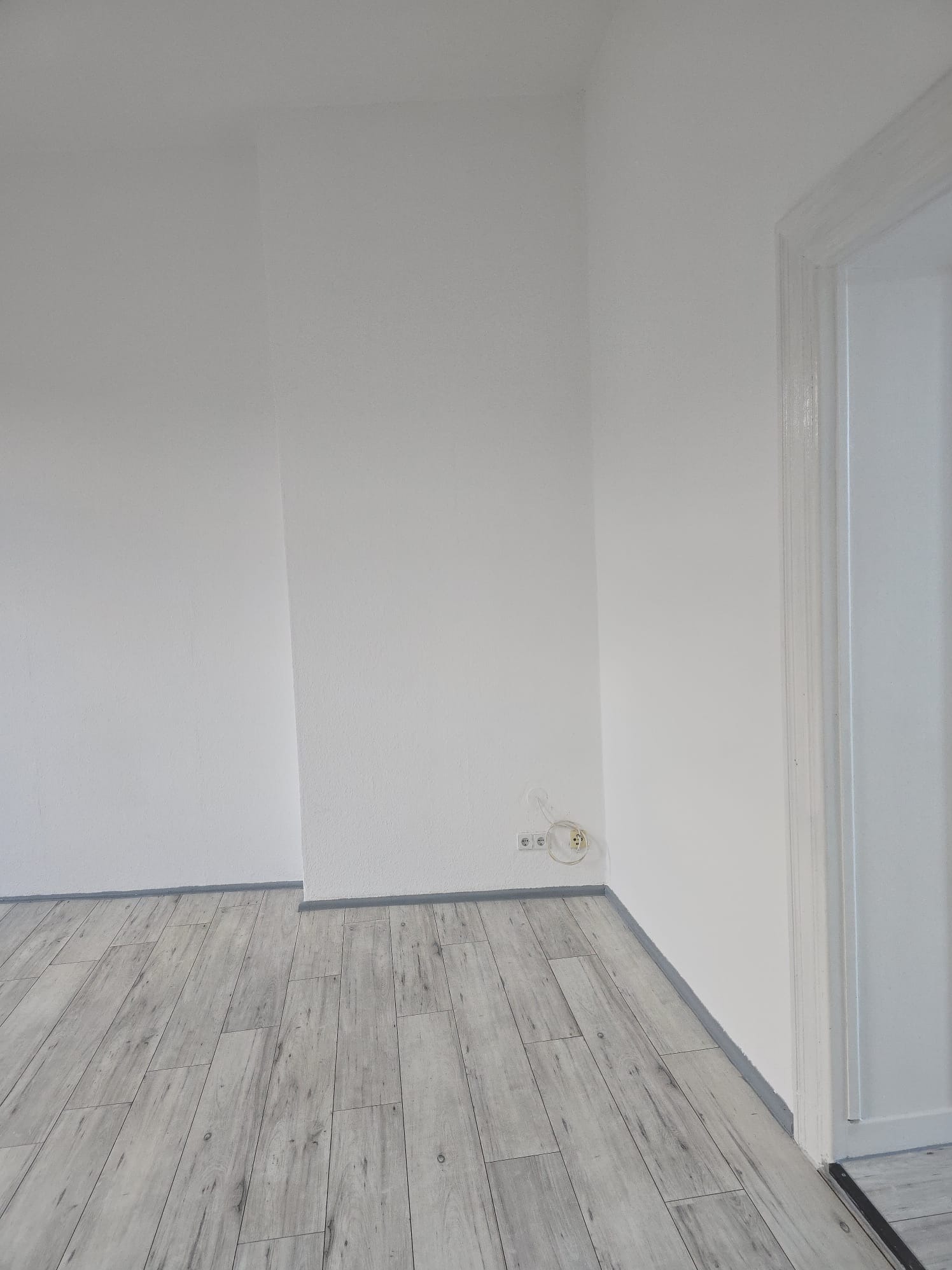 Jasne, odnowione wnętrze: świeżo pomalowane ściany w odcieniach bieli, szara podłoga imitująca drewno, widoczne gniazdka elektryczne i fragment białych drzwi.