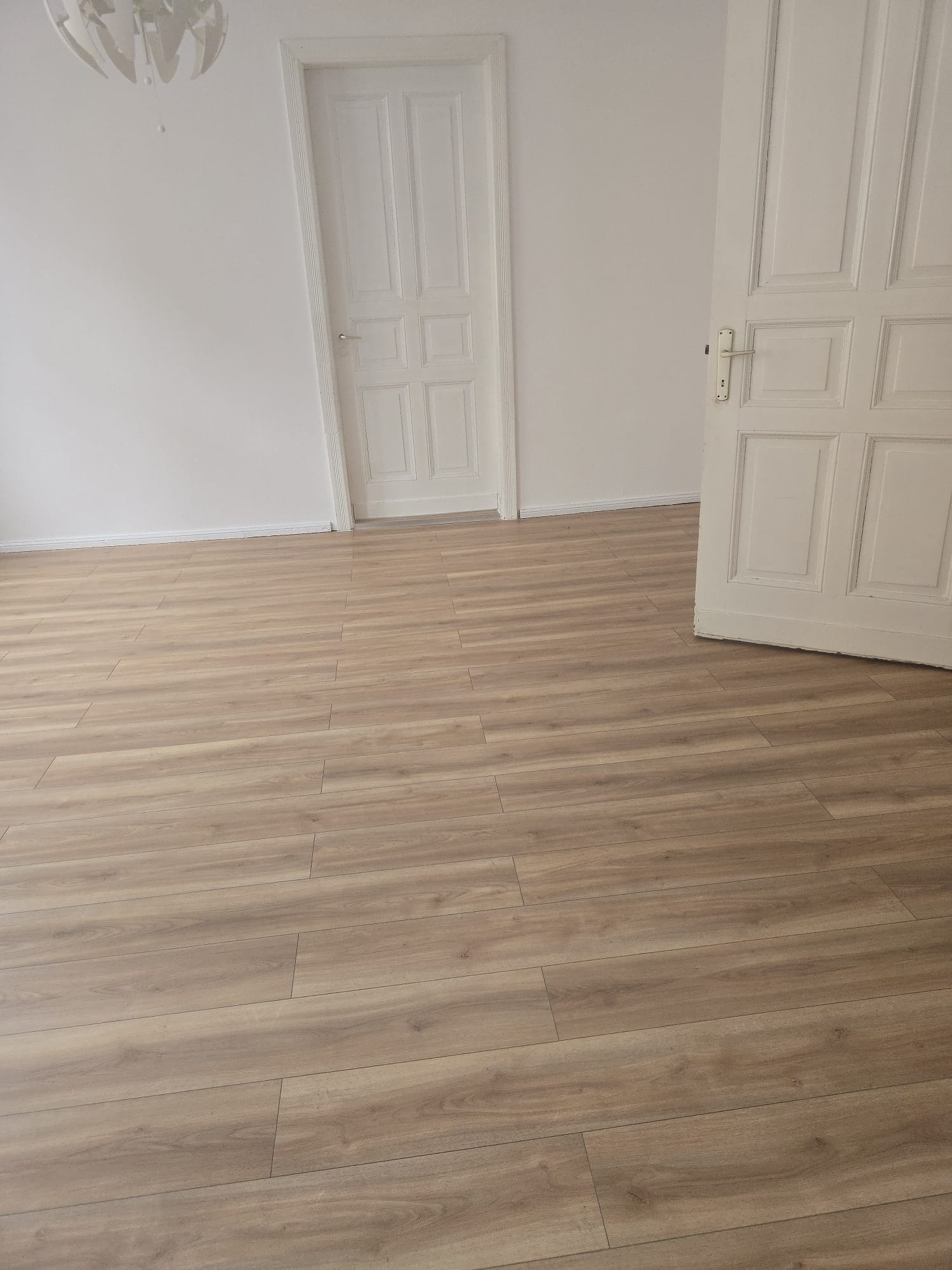 Jasna podłoga z paneli drewnopodobnych w pustym pokoju z białymi drzwiami. Widok na ułożone panele podłogowe z wyraźną strukturą drewna i otwarte drzwi.