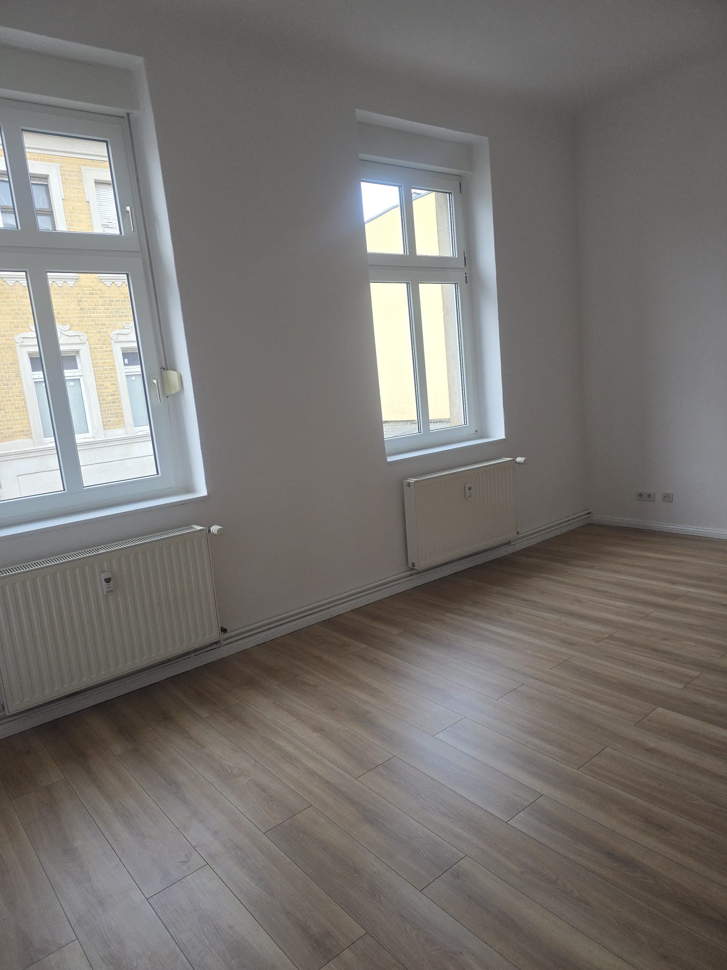 Jasne panele podłogowe w pustym pokoju z dwoma oknami i grzejnikami. Świeżo pomalowane ściany na biało. Widok na budynek z żółtej cegły za oknem.