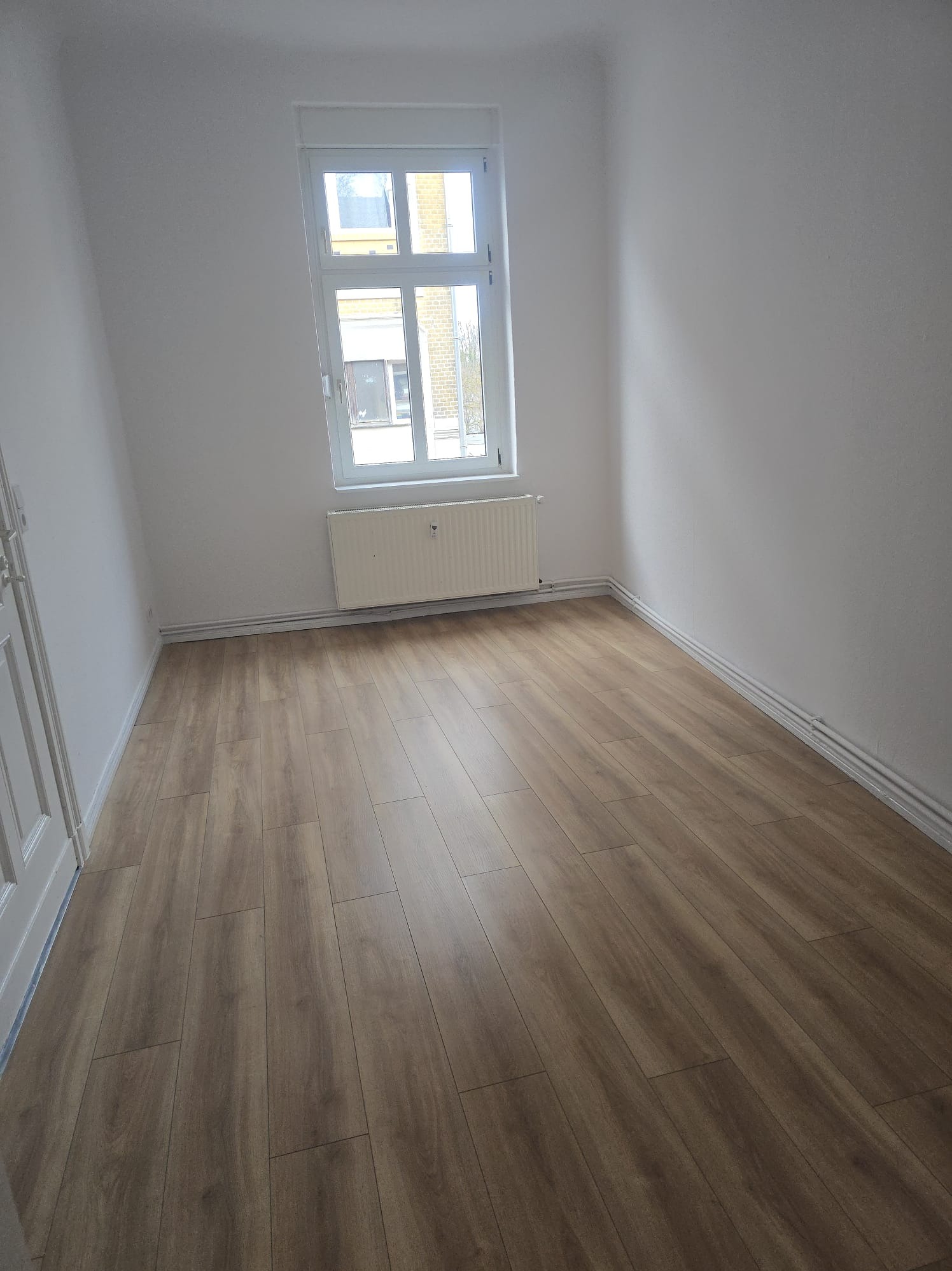 Jasne panele podłogowe w pustym, odświeżonym pokoju z białymi ścianami i oknem. Widoczne rury grzewcze przy podłodze. Minimalistyczny wystrój, świeżo po remoncie.