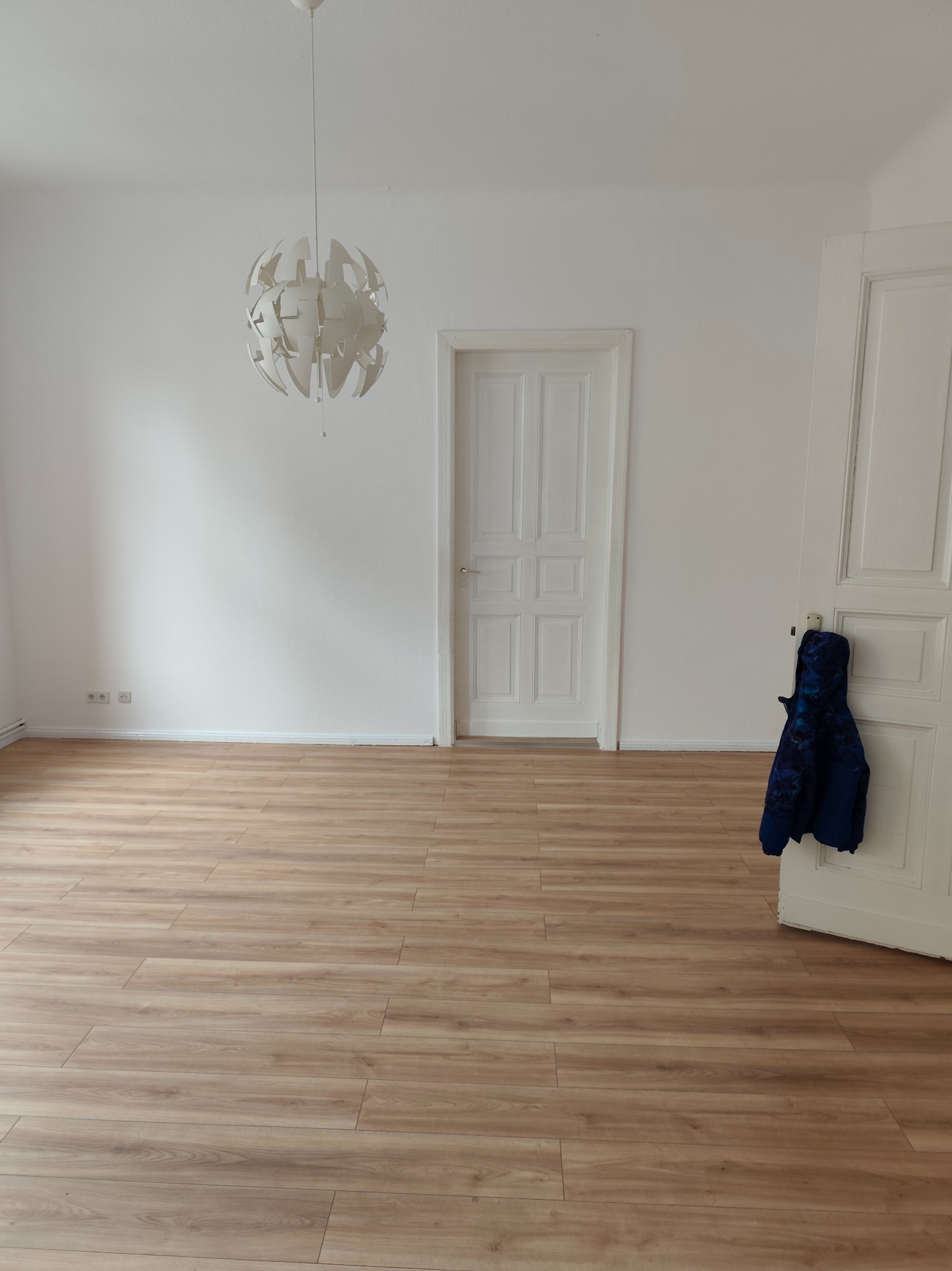 Jasne panele podłogowe w pustym pokoju z białymi ścianami i drzwiami. Nowoczesna lampa wisząca. Niebieska kurtka wisi na klamce drzwi.