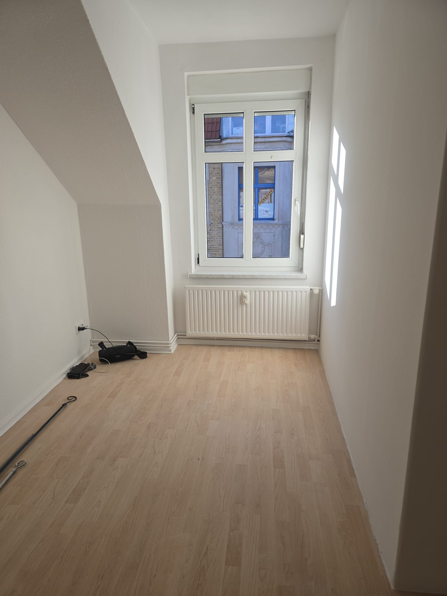 Jasne, puste poddasze z oknem i drewnianą podłogą. Świeżo pomalowane ściany, grzejnik pod oknem. Kabel zasilający i rura leżą na podłodze. Widok na budynek zza okna.