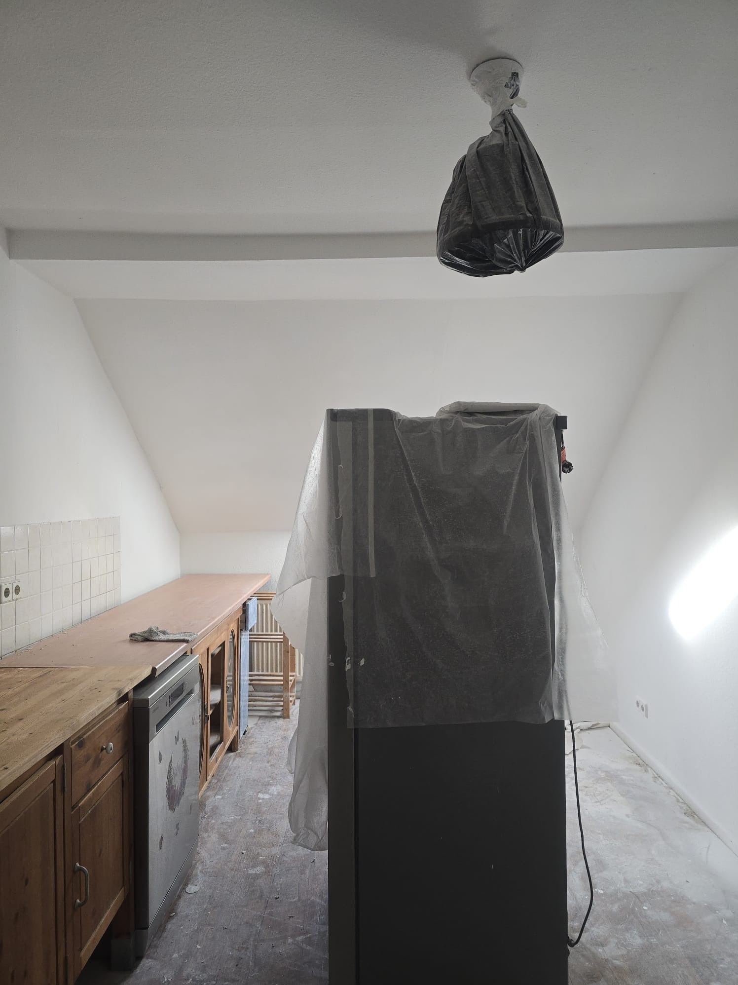 Kuchnia w trakcie remontu: meble zabezpieczone folią, sufit i ściany świeżo pomalowane na biało, podłoga częściowo zakryta. Lampa owinięta folią ochronną.
