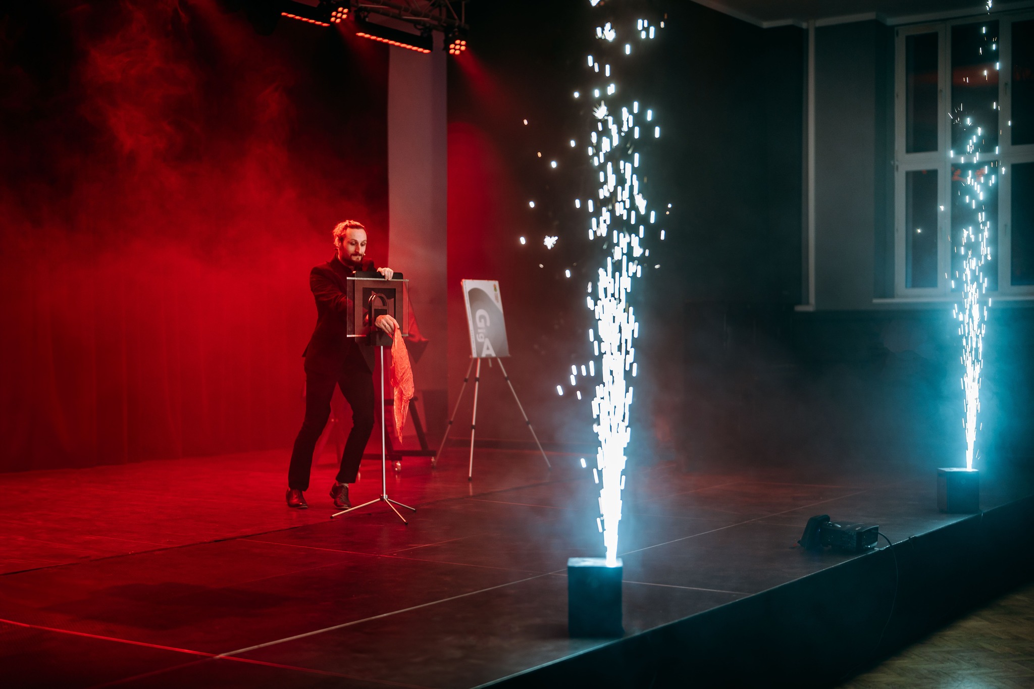 Iluzjonista w trakcie pokazu z rekwizytami i efektami pirotechnicznymi na scenie w czerwono-niebieskim oświetleniu. W tle sztaluga z logotypem.