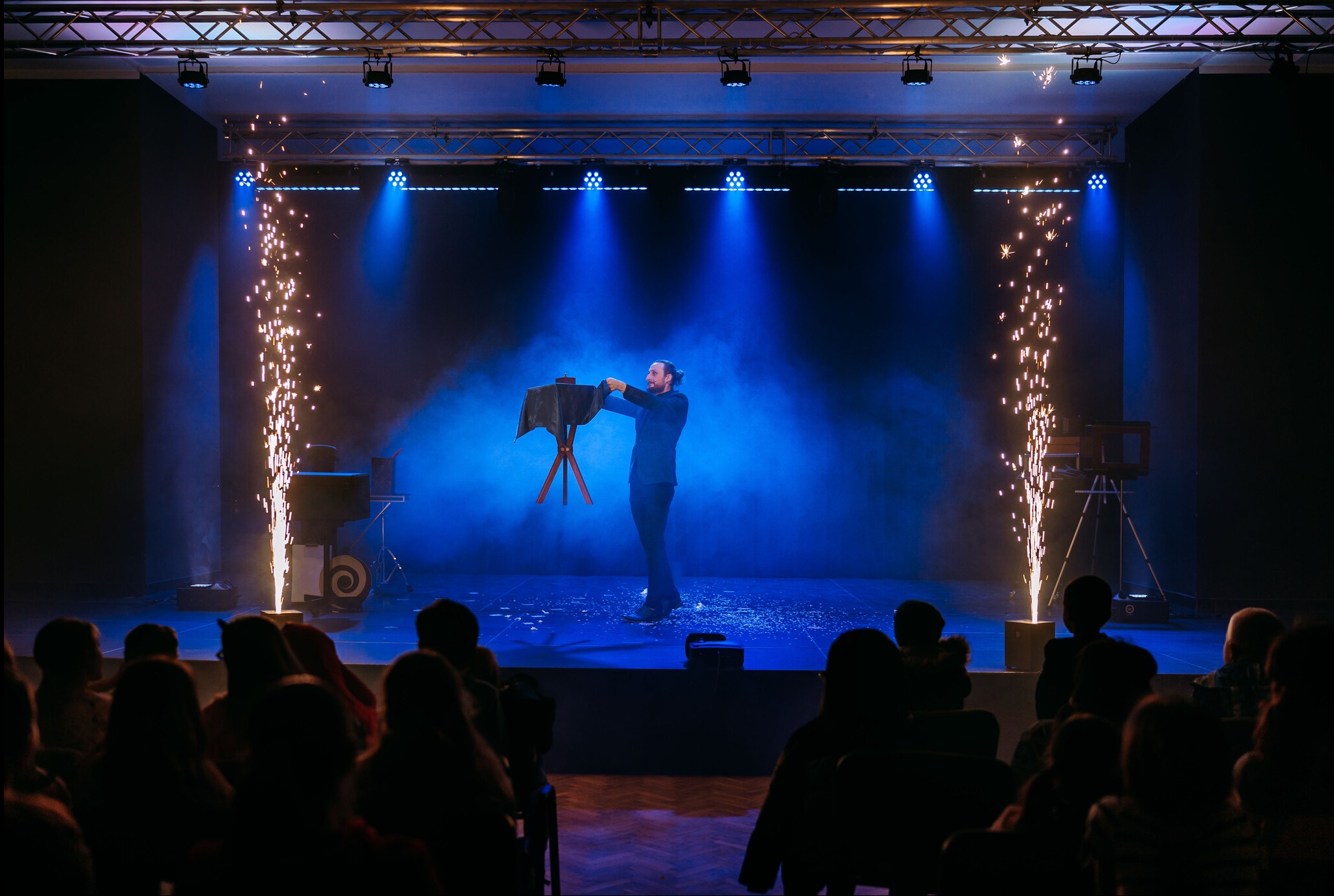 Iluzjonista na scenie, w tle niebieskie światła i efekty pirotechniczne, widownia w cieniu. Magik prezentuje trik z użyciem stolika i czarnej tkaniny.