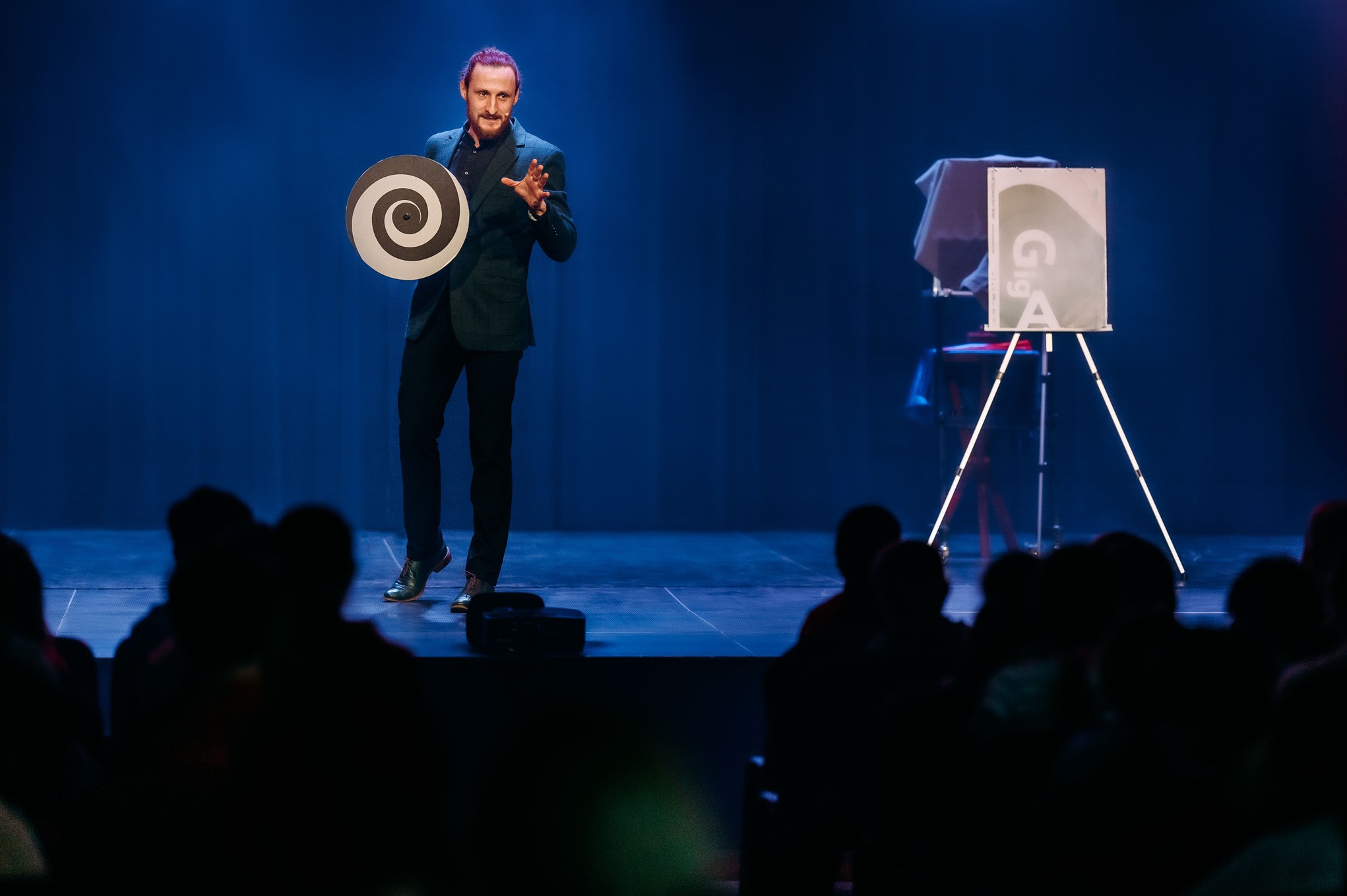 Iluzjonista na scenie trzymający rekwizyt z hipnotyczną spiralą, w tle publiczność i stolik z przykrytym przedmiotem. Scena oświetlona niebieskim światłem.