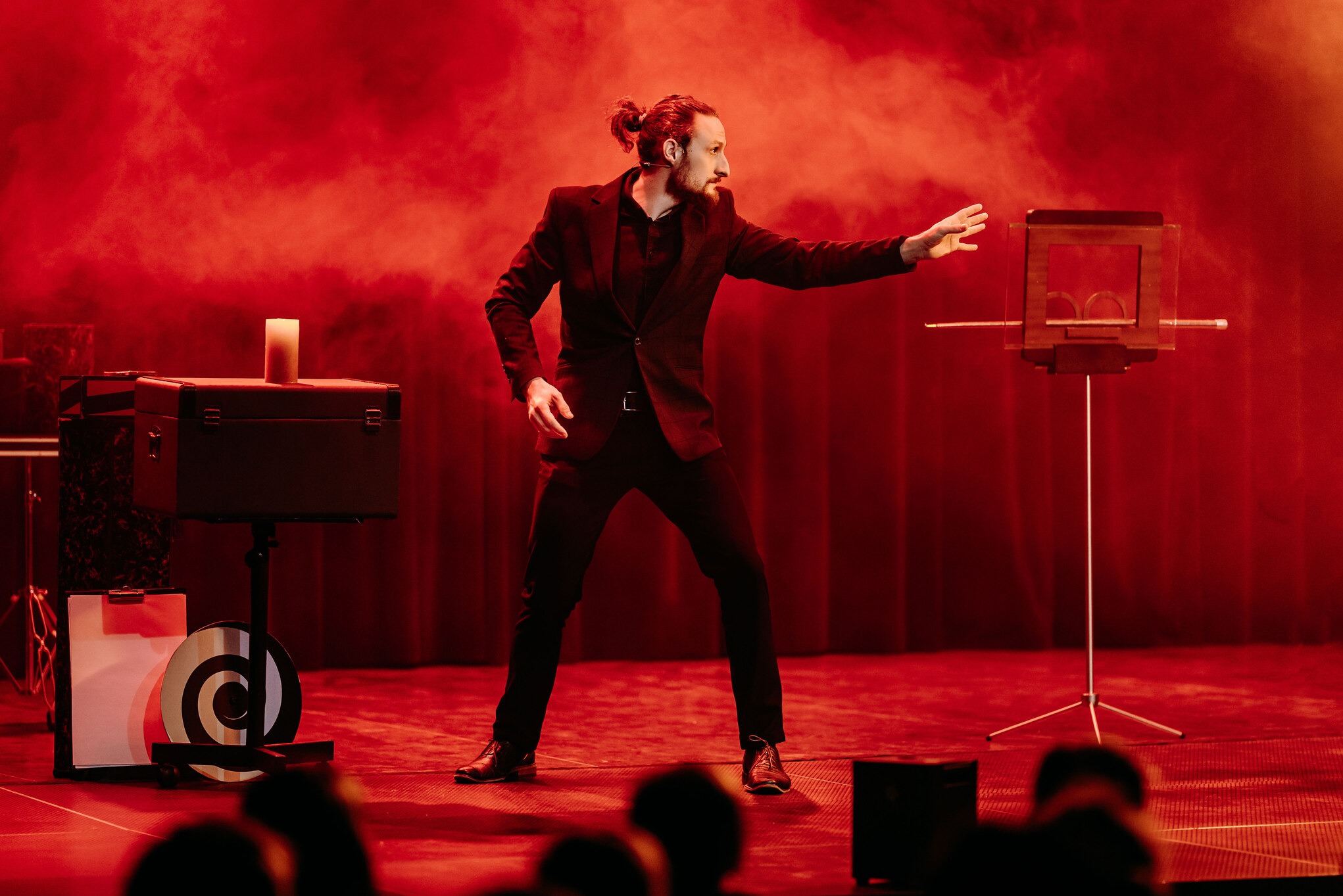 Iluzjonista w czarnym garniturze na scenie w czerwonym oświetleniu, wykonujący trik z rekwizytem przypominającym ramkę. W tle skrzynia i inne akcesoria.