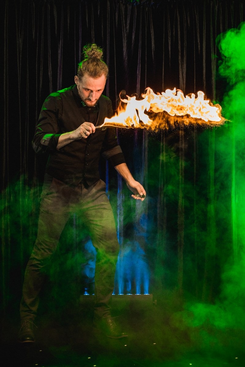 Iluzjonista z płonącą dłonią na scenie, w otoczeniu zielonego dymu i ciemnych zasłon. Efektowny pokaz z ogniem, element występu artystycznego.