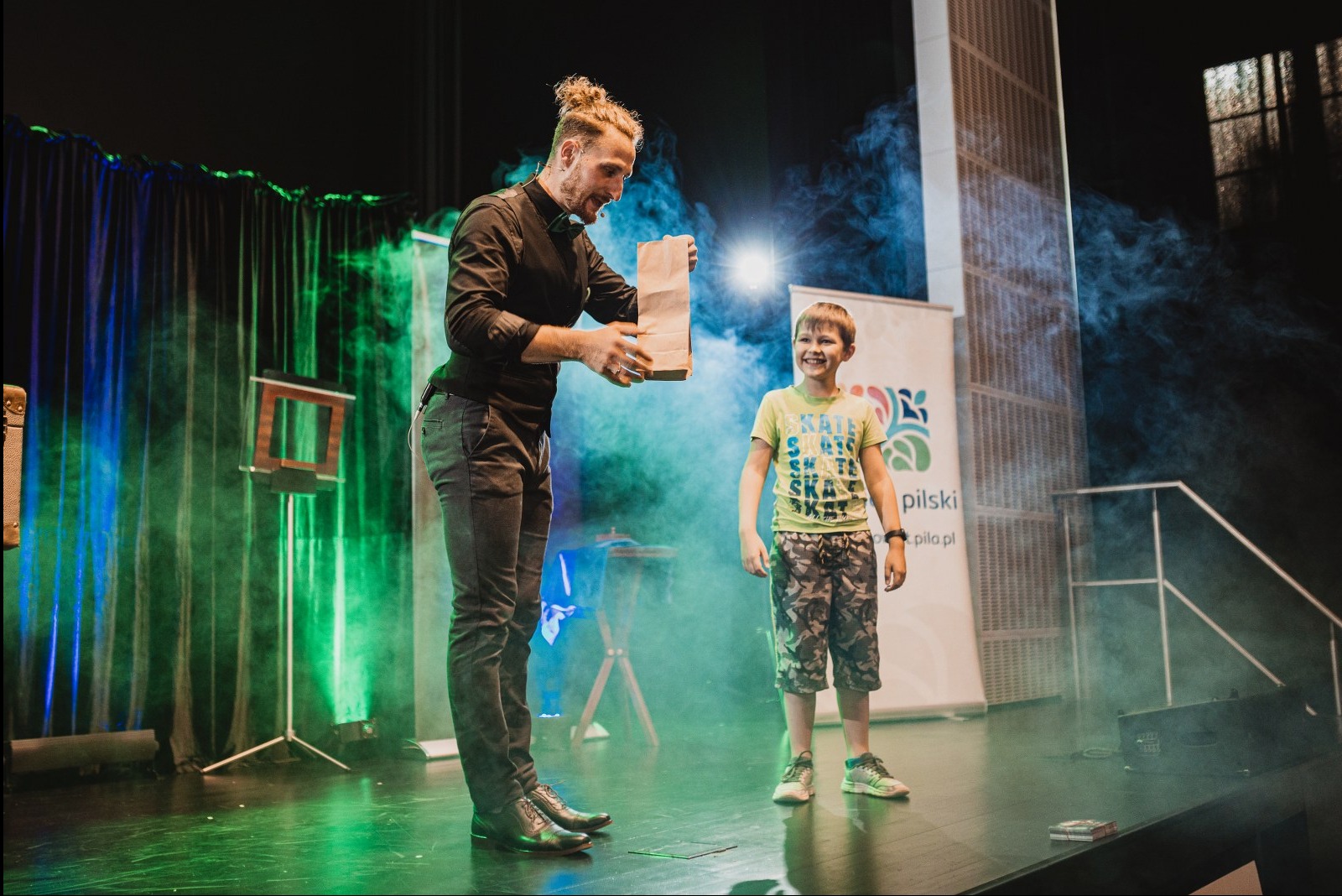Iluzjonista w czarnej koszuli prezentuje trik z papierową torbą uśmiechniętemu chłopcu na scenie w oparach dymu i zielono-niebieskich świateł.