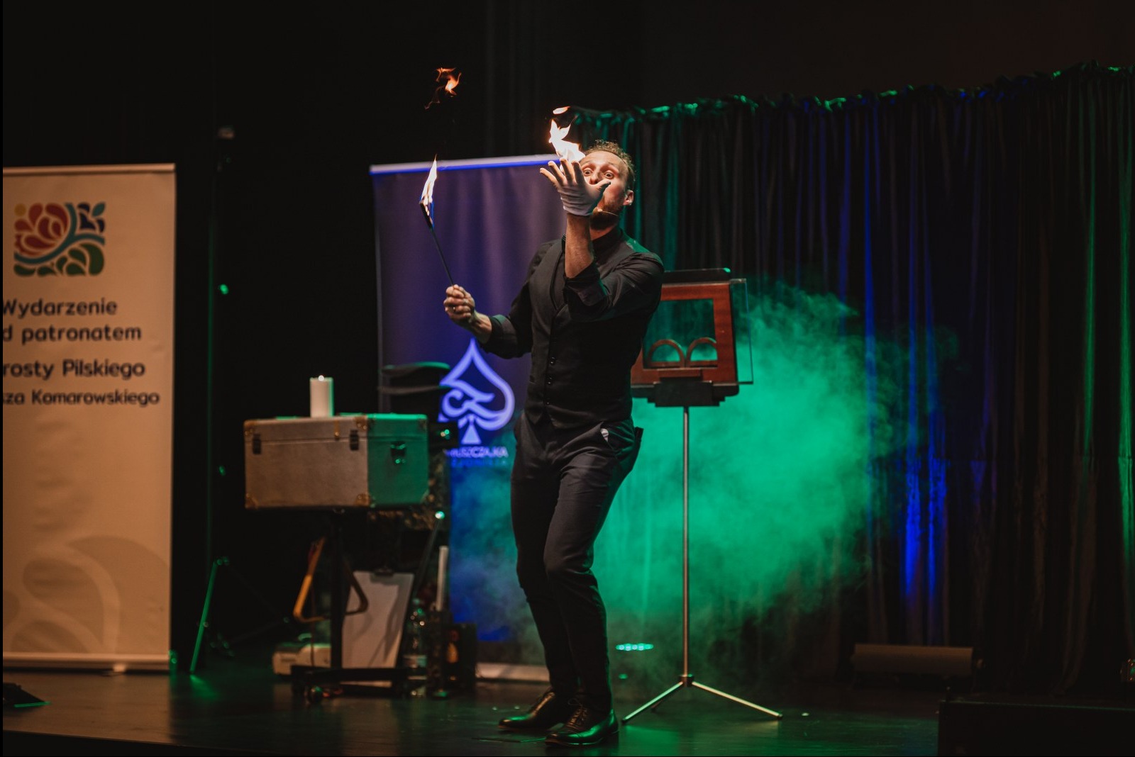 Iluzjonista w czarnym stroju żongluje ogniem na scenie, w tle rekwizyty i zielone światło. Dynamiczne ujęcie z występu.