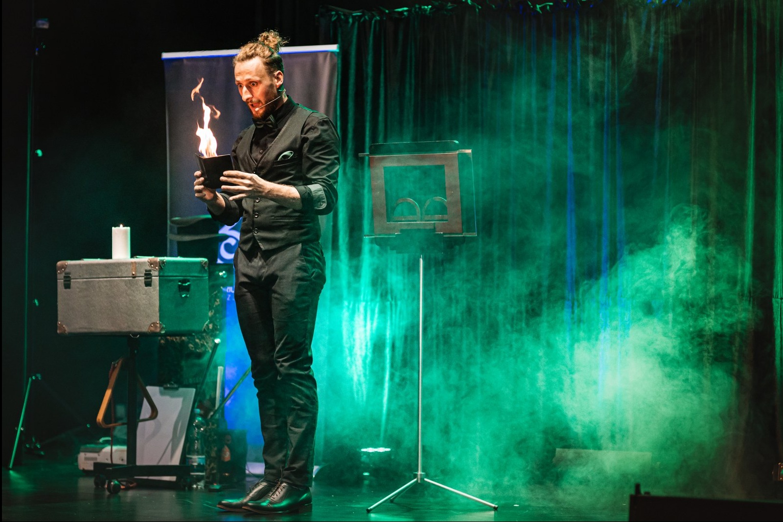 Iluzjonista w czarnym stroju prezentuje płonący rekwizyt na scenie z zielonymi światłami i dymem. W tle skrzynia i stojak z logo.