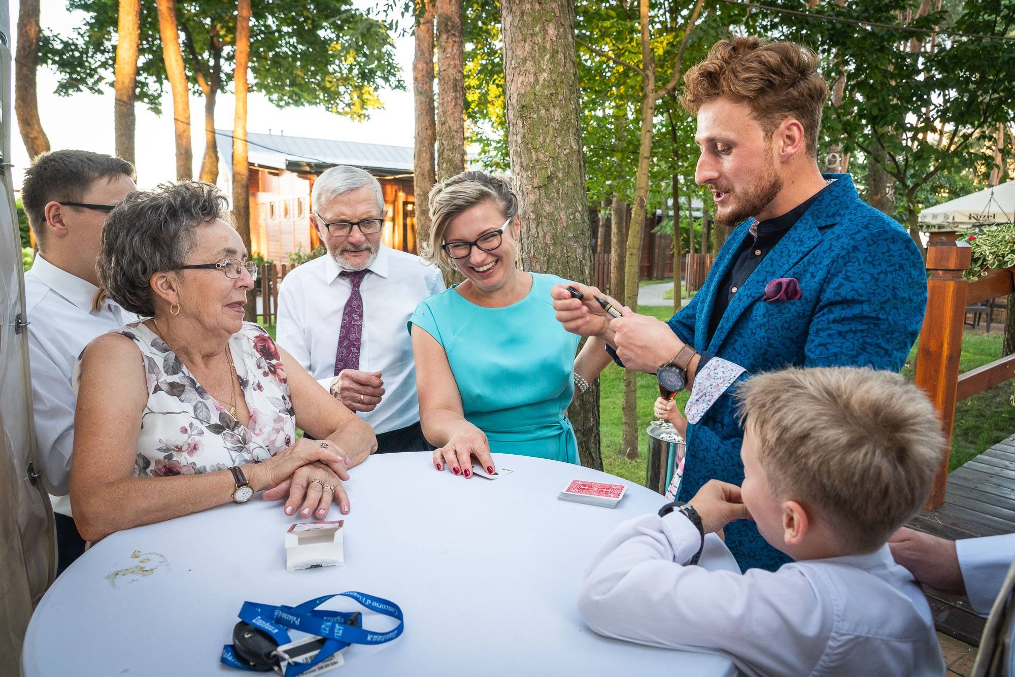 Iluzjonista w błękitnej marynarce prezentuje trik z kartami grupie widzów przy okrągłym stole na zewnątrz, w tle drzewa i budynek.
