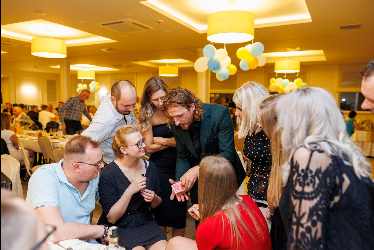 Iluzjonista w zielonym smokingu prezentuje trik z kartami grupie widzów w sali balowej z dekoracjami balonowymi i oświetleniem punktowym.