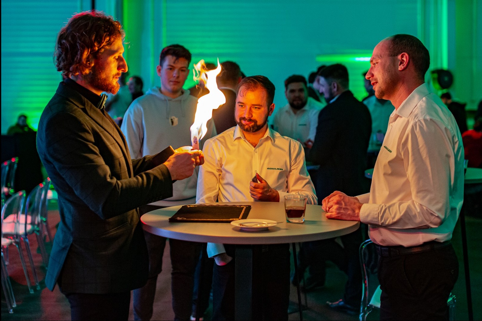 Iluzjonista prezentuje trik z ogniem przy stoliku podczas eventu firmowego. W tle widoczni goście i zielone oświetlenie.