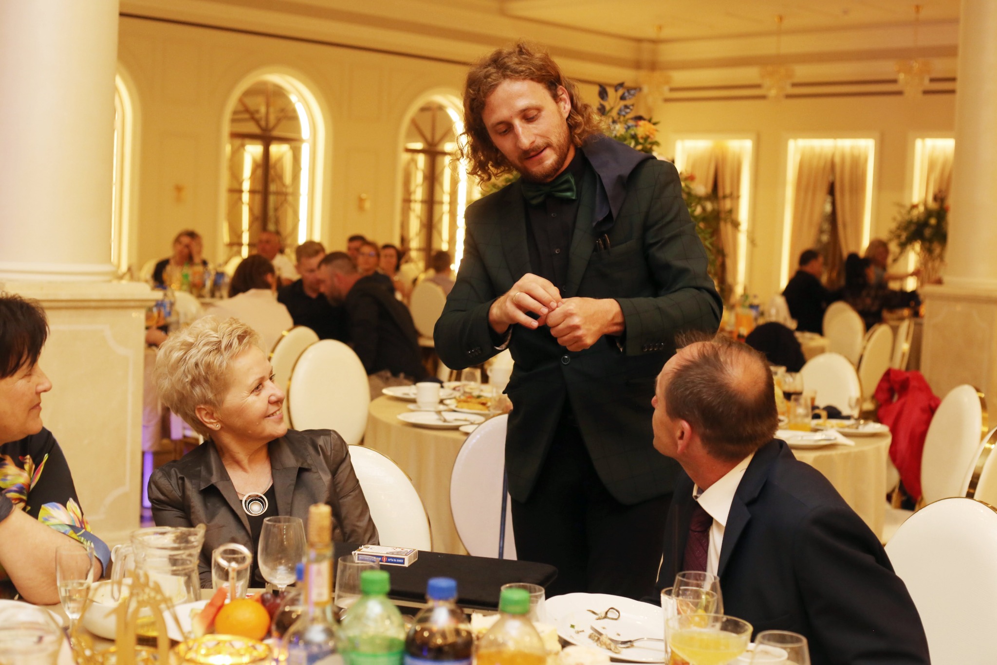 Iluzjonista w zielonym smokingu prezentuje trik z kartami przy stole podczas eleganckiego przyjęcia. W tle goście i dekoracje.