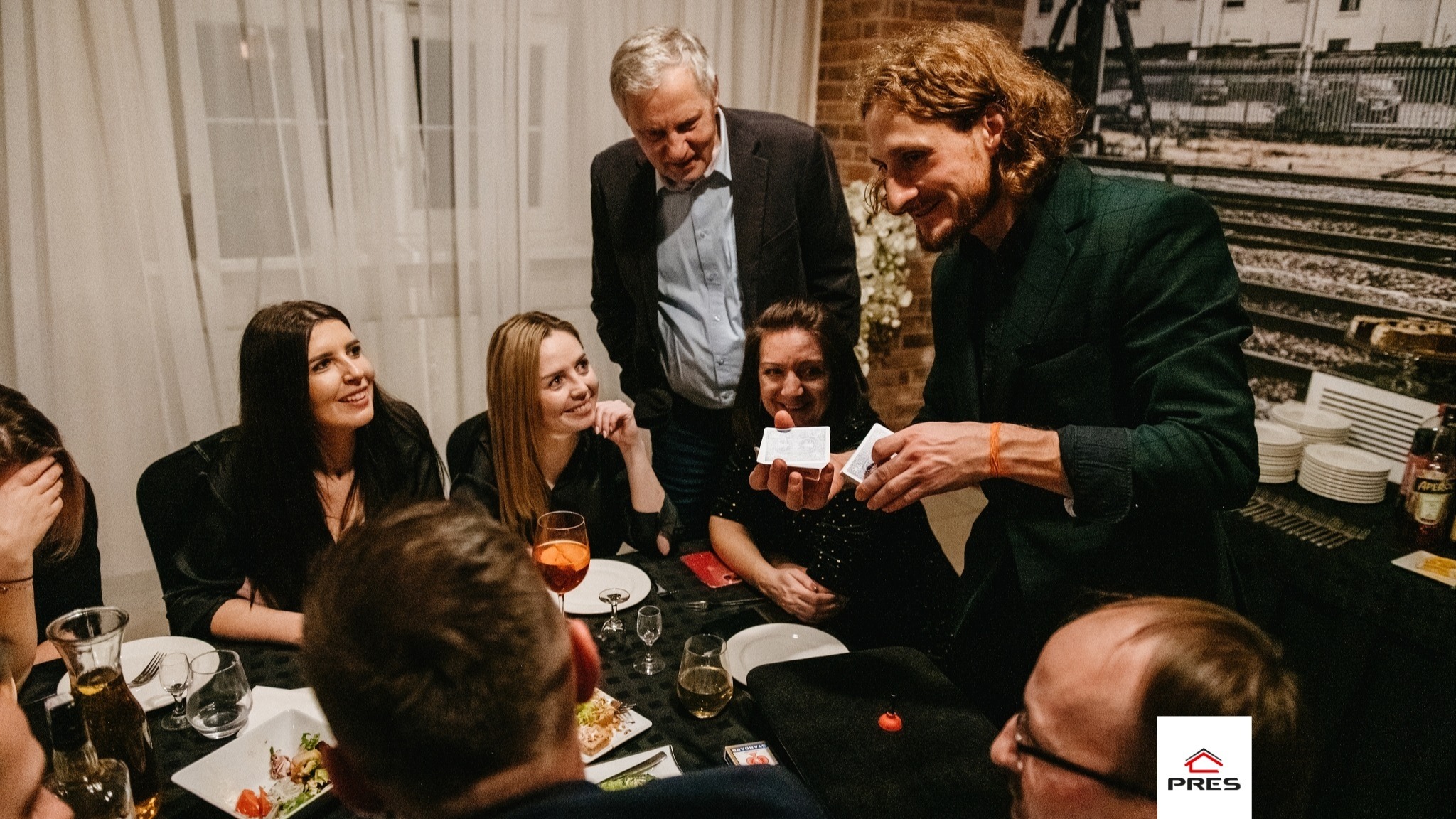 Iluzjonista prezentuje trik z kartami grupie osób siedzących przy stole podczas kameralnego spotkania. W tle widoczna dekoracja ściany z motywem kolejowym.