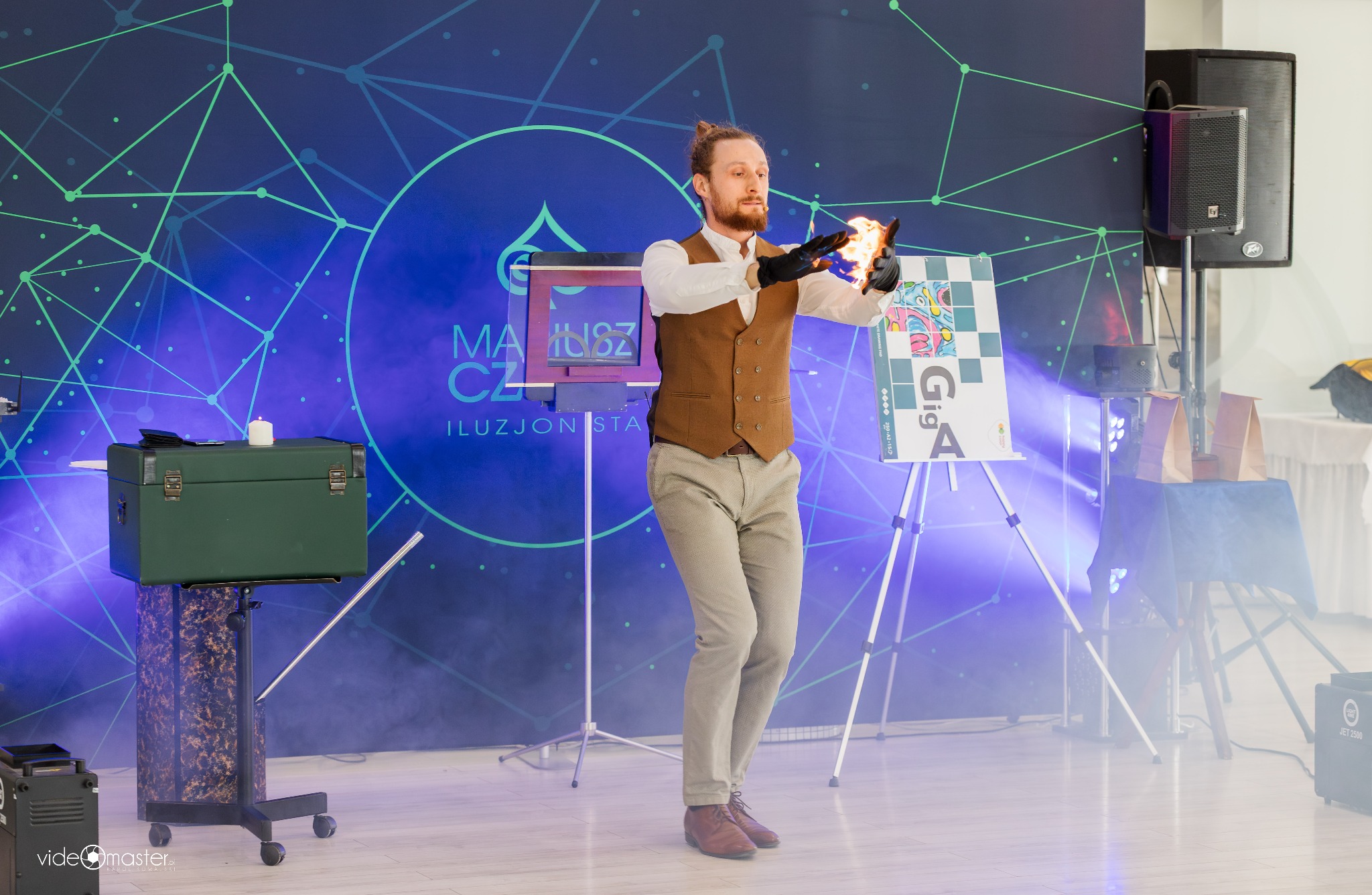 Iluzjonista w Łubiance prezentuje trik z ogniem na tle dekoracji scenicznej. Widoczny rekwizyt, skrzynia i oświetlenie sceniczne.