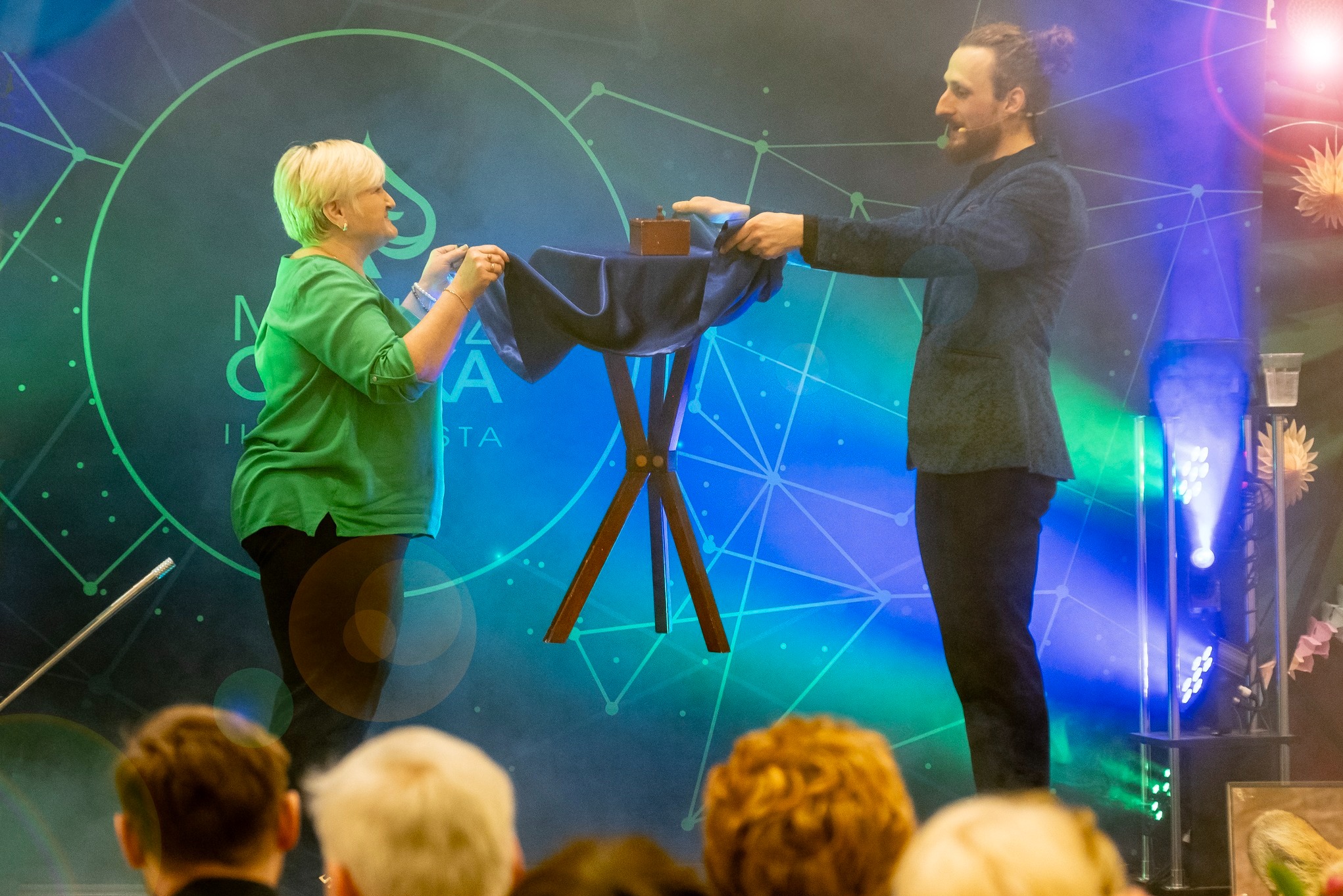 Iluzjonista prezentuje trik z drewnianym pudełkiem na stoliku, asystentka pomaga w pokazie na tle sceny z oświetleniem i publicznością.