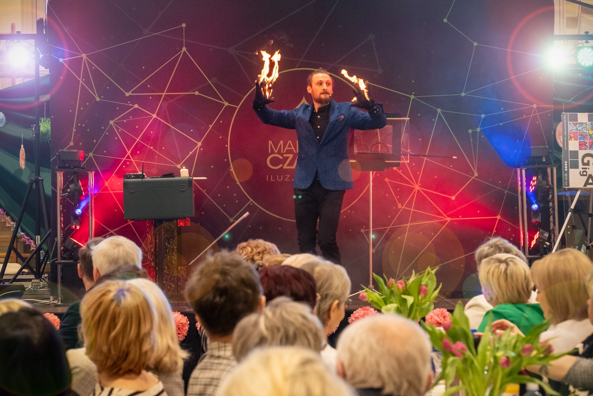 Iluzjonista w niebieskiej marynarce prezentuje płonące dłonie na tle sceny z geometrycznym wzorem, widziany z perspektywy publiczności.