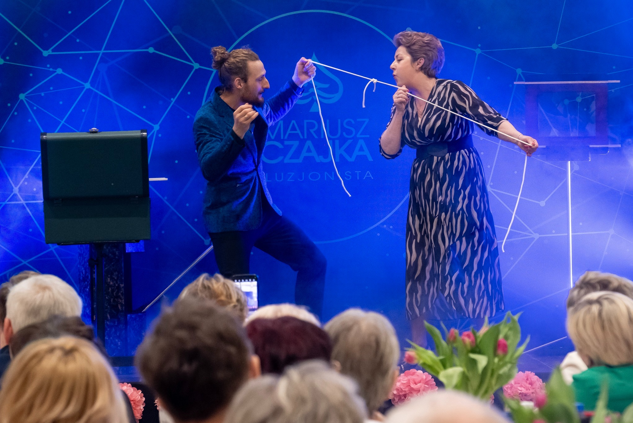 Iluzjonista Mariusz Czajka prezentuje trik z liną podczas występu na scenie z niebieskim tłem. Widzowie obserwują z zaciekawieniem.