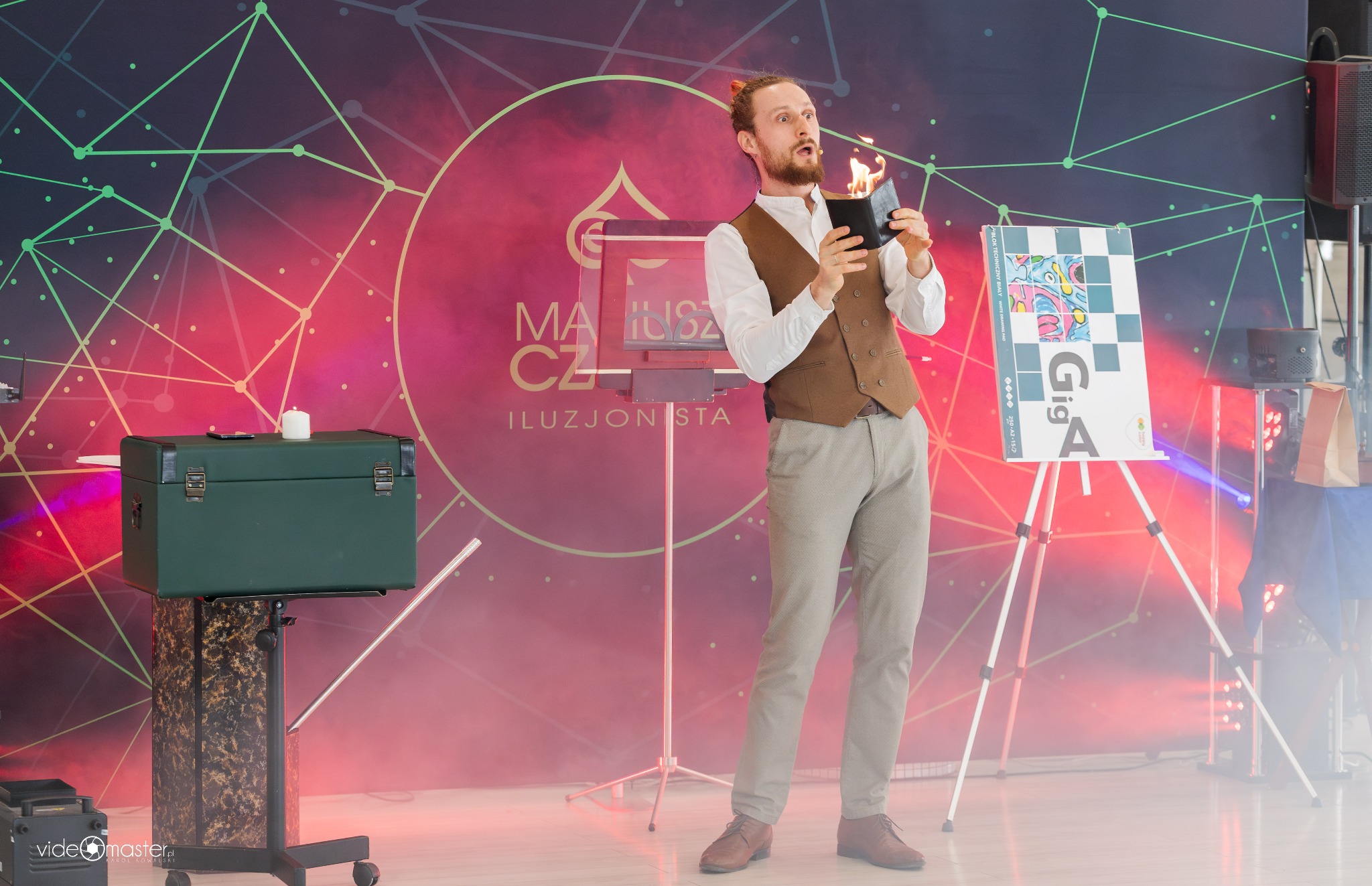 Iluzjonista Mariusz Cz. w trakcie pokazu z płonącym portfelem na tle grafiki z logo. Na scenie rekwizyty: kufer, stolik, sztaluga. Czerwone oświetlenie sceniczne.