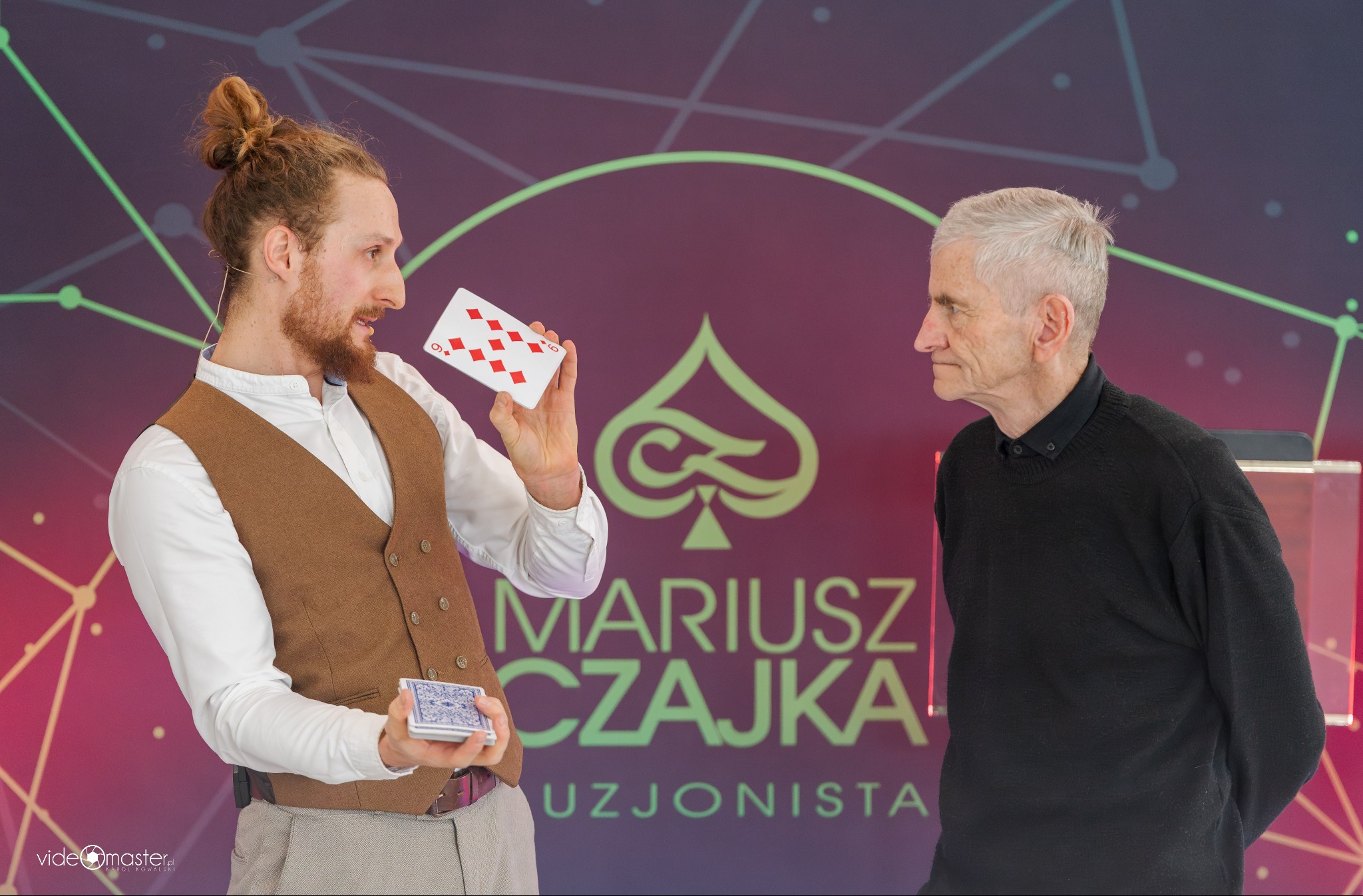 Iluzjonista prezentuje trik z kartami przed widzem na tle sceny z logo. Mężczyzna w kamizelce trzyma talię kart i jedną kartę z numerem 9 w romby.