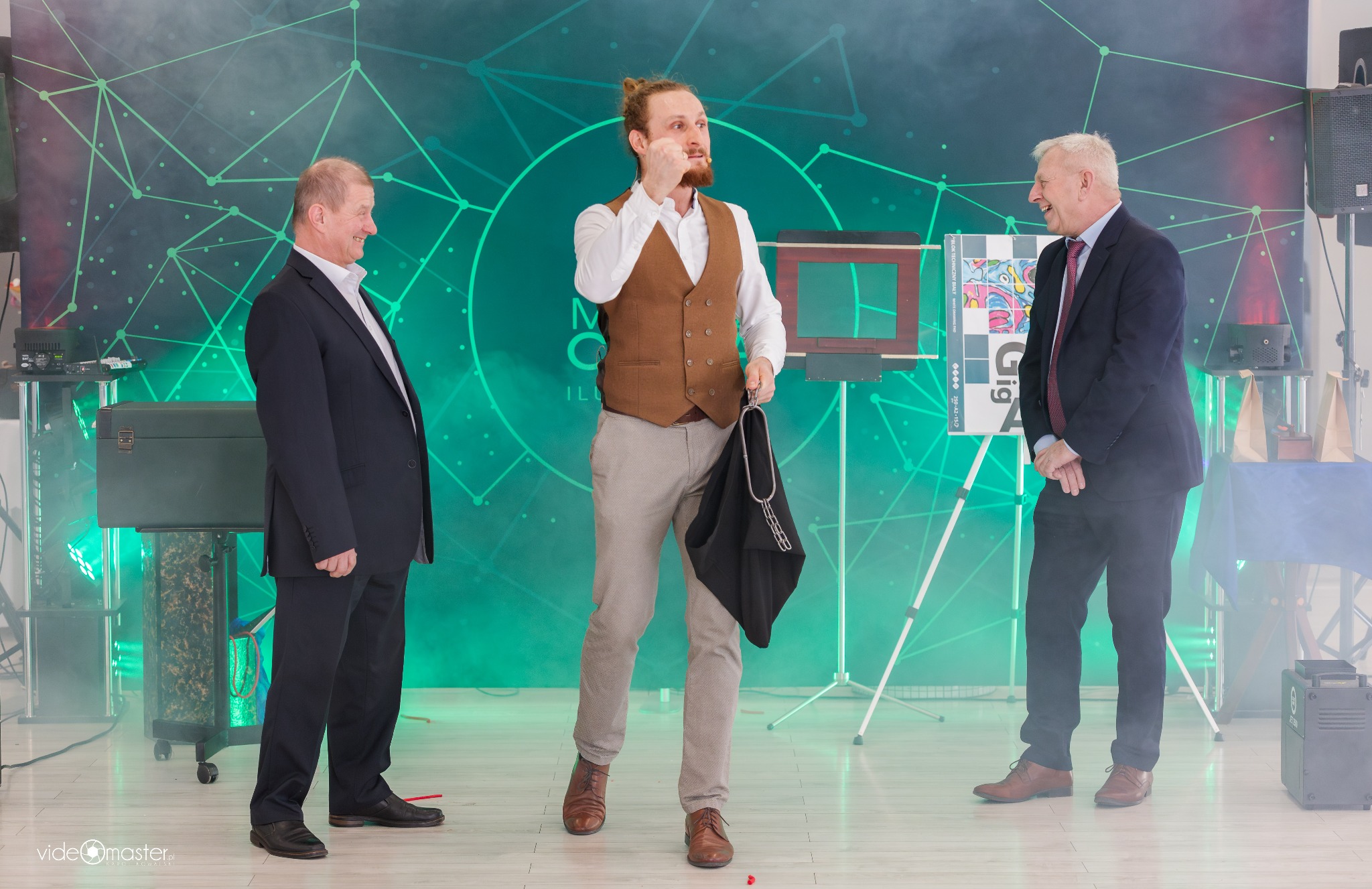 Iluzjonista w brązowej kamizelce prezentuje trik z czarnym workiem na tle zielonej scenografii, otoczony uśmiechniętymi widzami w garniturach.