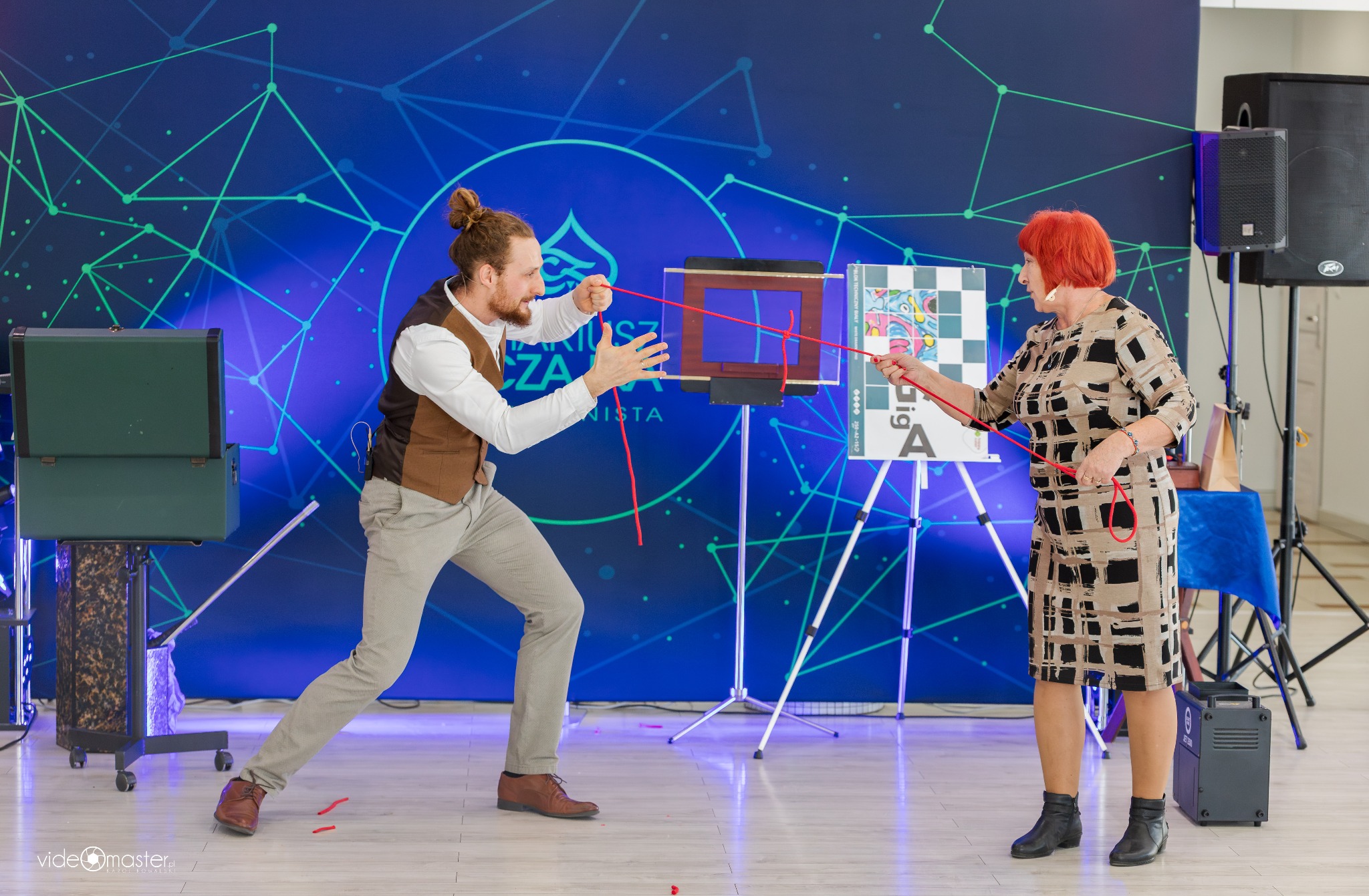 Iluzjonista z brodą i kobieta z czerwoną peruką wykonują trik z czerwoną liną na tle niebieskiej sceny z geometrycznym wzorem. Widoczne rekwizyty i sprzęt nagłośnieniowy.