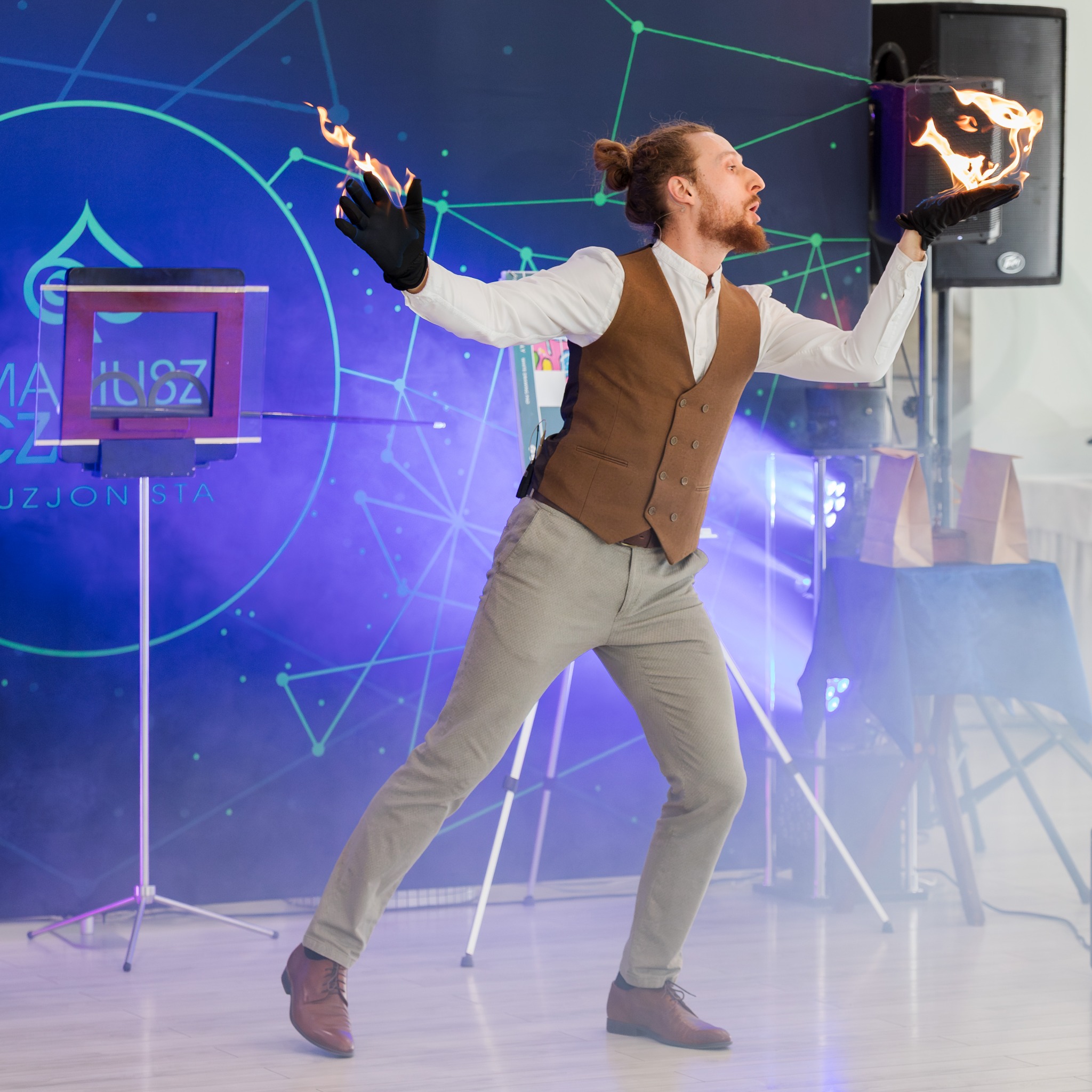 Iluzjonista w brązowej kamizelce prezentuje płomienie na dłoniach podczas występu na tle niebieskiego ekranu z geometrycznym wzorem i sprzętem.