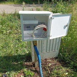 Rafał Cegliński - Otwarta biała skrzynka elektryczna z bezpiecznikami i zwiniętym kablem z czerwoną wtyczką siłową. Niebieski peszel wychodzi z ziemi, na tle zielonej trawy i drogi.