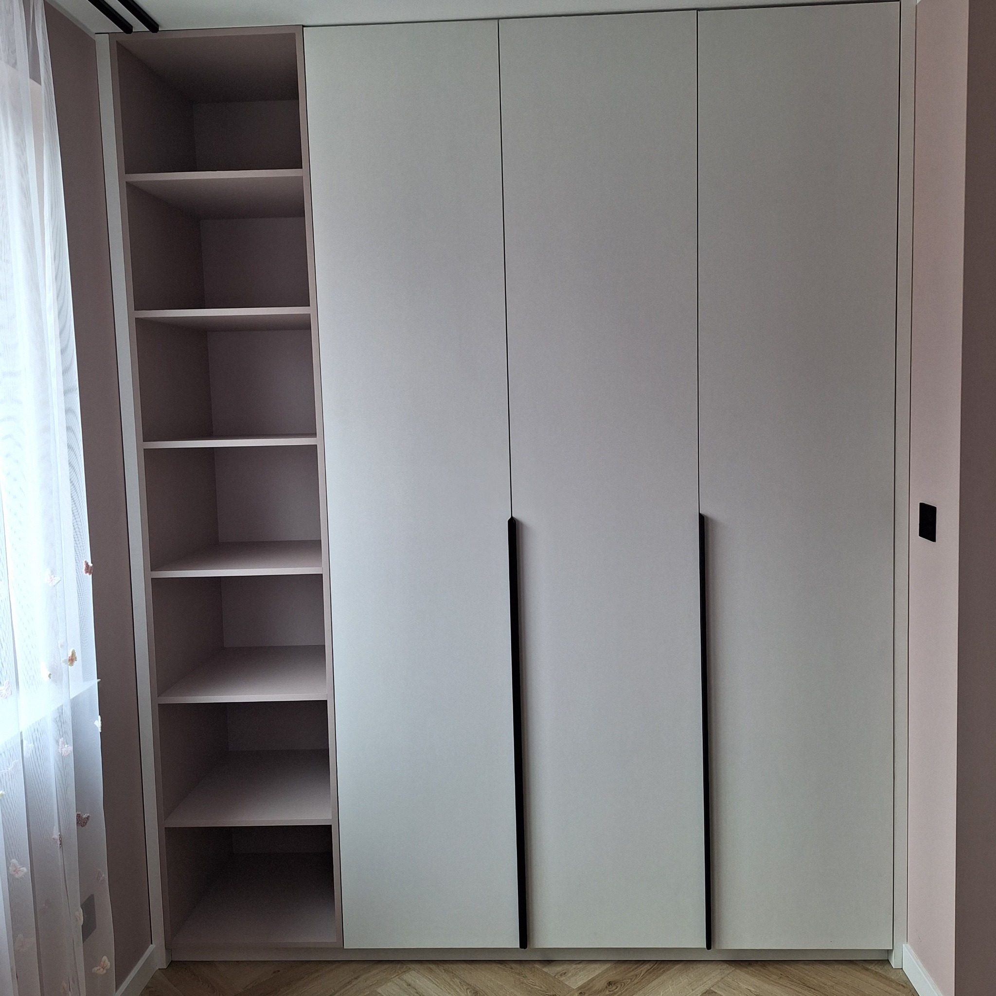 Jasna szafa z trzema drzwiami i otwartą półką z boku, minimalistyczne czarne uchwyty, parkiet w jodełkę, delikatna firanka w tle.