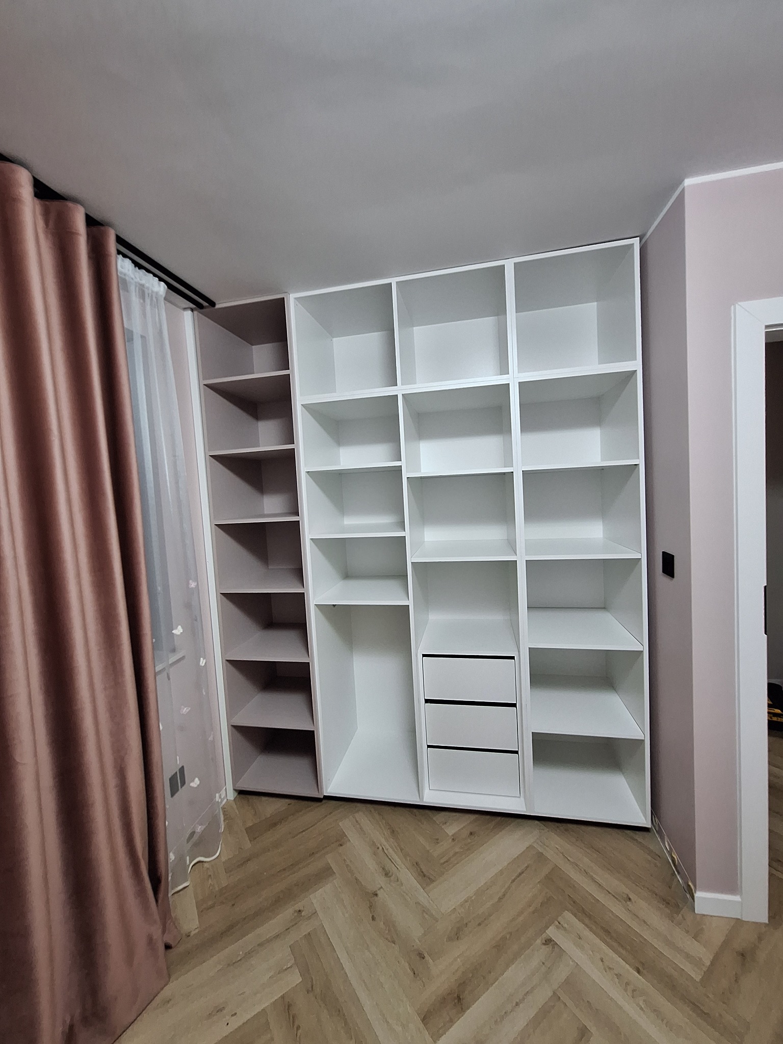 Biała szafa wnękowa z szufladami i półkami, część regału w kolorze taupe, parkiet w jodełkę, zasłona w odcieniach brązu, ściana w kolorze pudrowego różu.