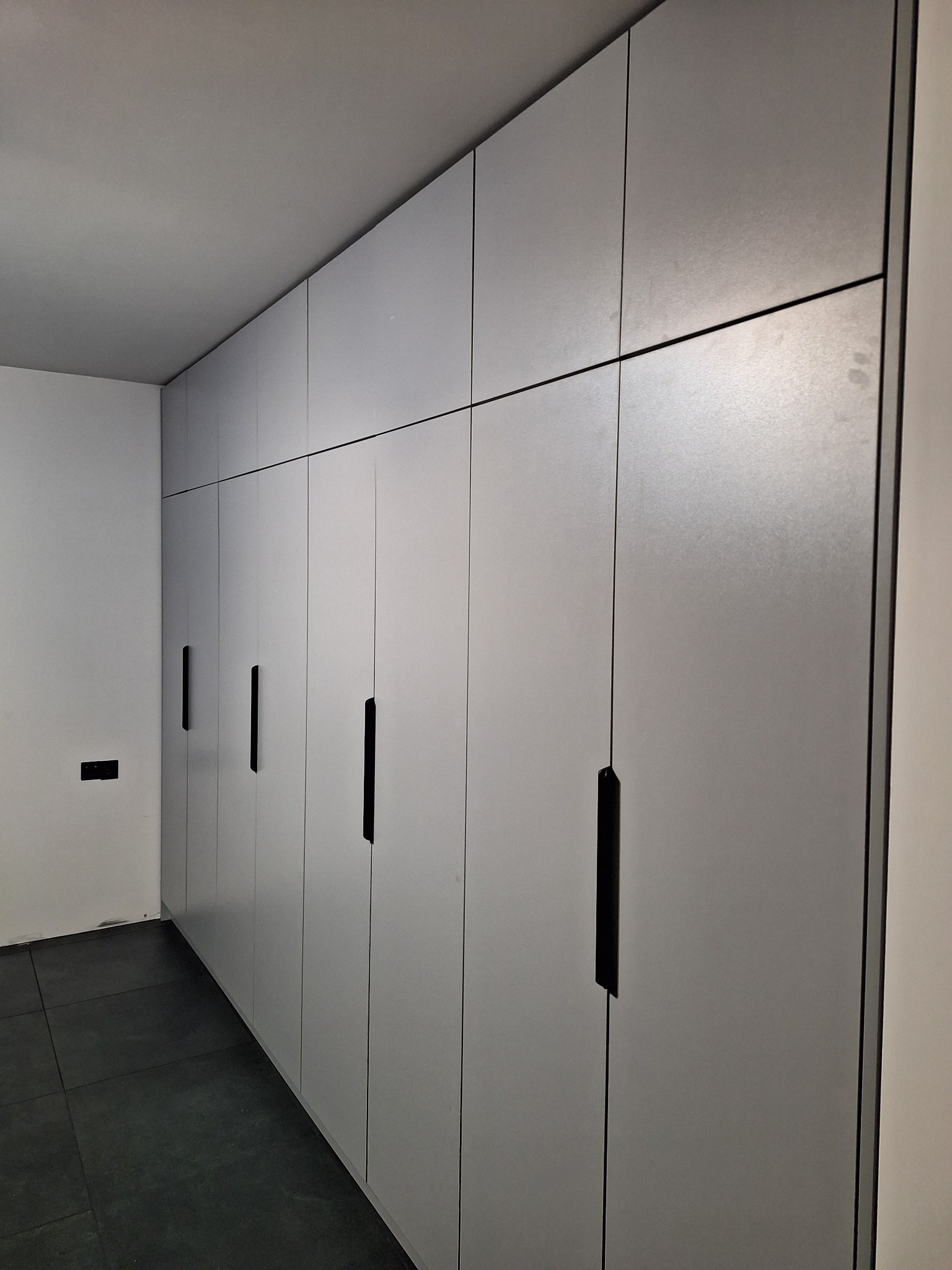 Długa, zabudowana szafa wnękowa z szarymi frontami i czarnymi, pionowymi uchwytami, dopasowana do wnętrza z ciemną podłogą. Minimalistyczny design.