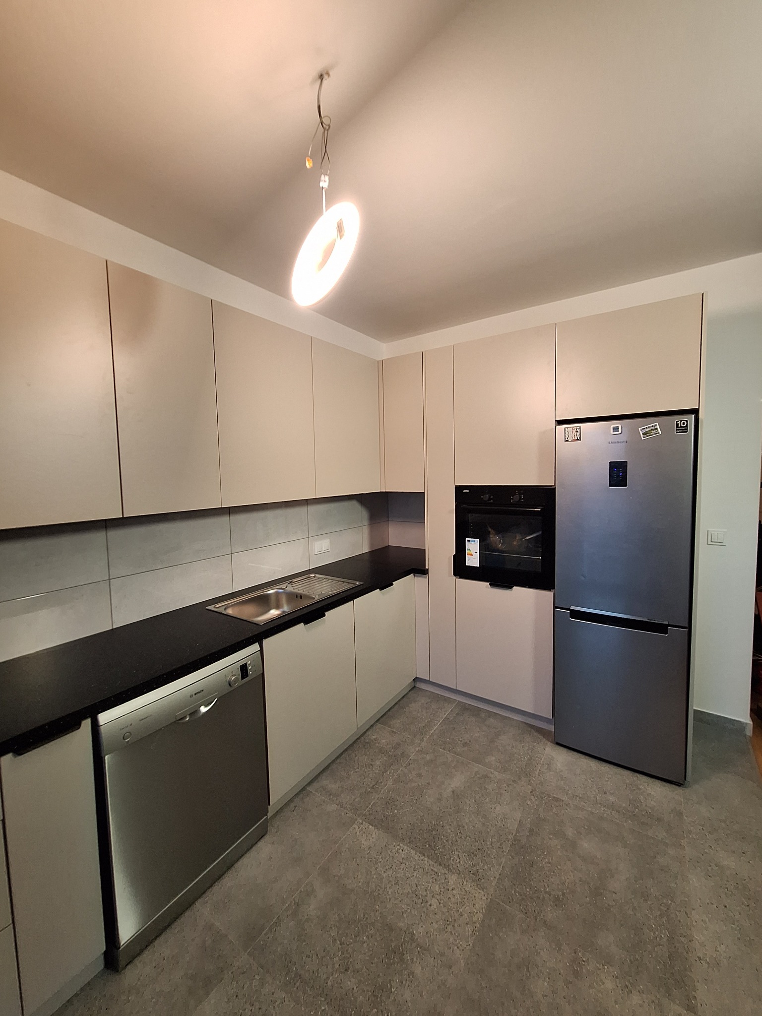 Jasna kuchnia na wymiar z szarymi płytkami i czarnym blatem. Zabudowa obejmuje piekarnik i lodówkę. Widoczna zmywarka i oświetlenie sufitowe.