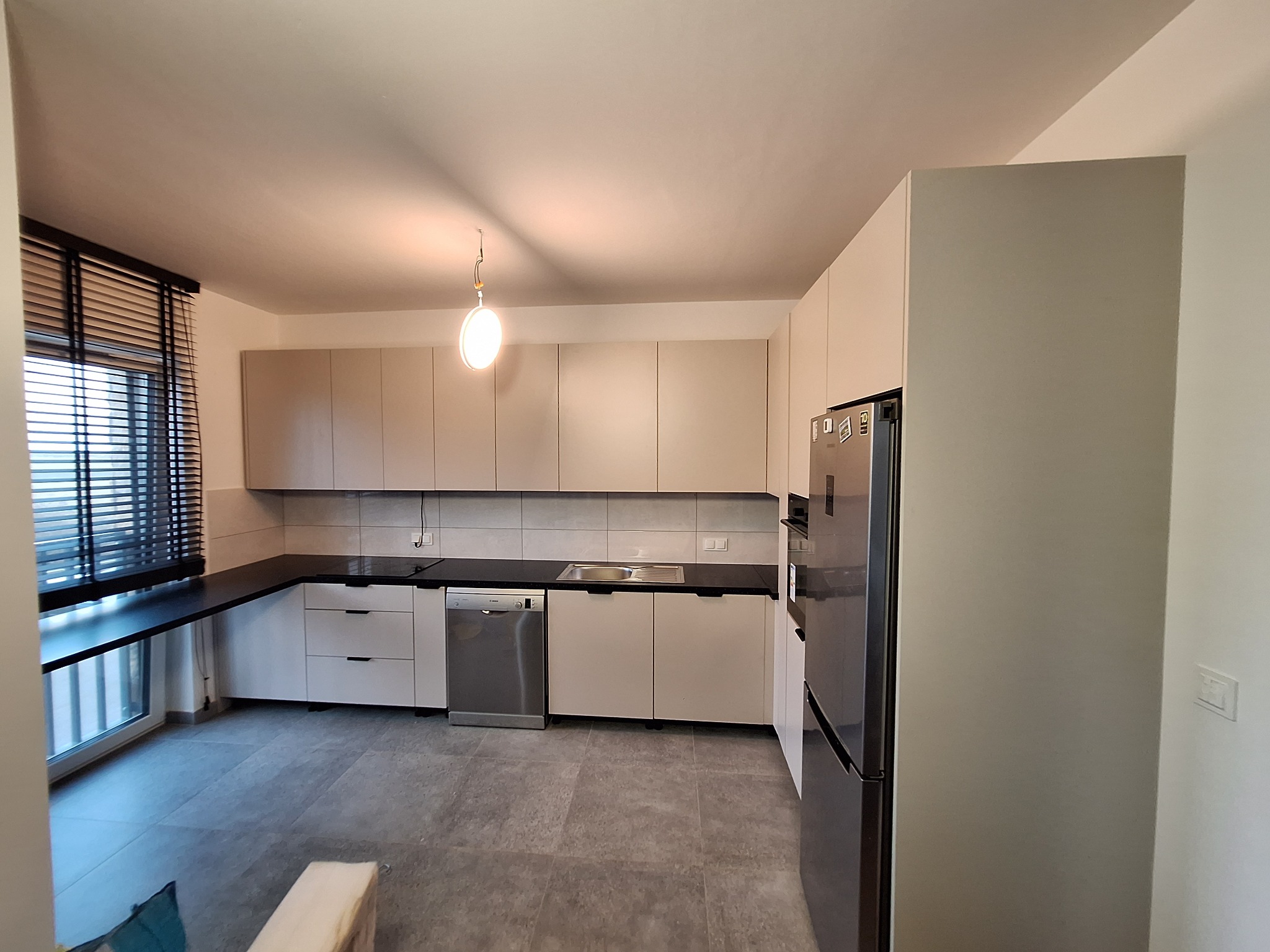 Jasna kuchnia z zabudową na wymiar, czarnym blatem i szarymi płytkami. Widoczne okno z żaluzjami i nowoczesna lampa sufitowa. Minimalistyczny design.