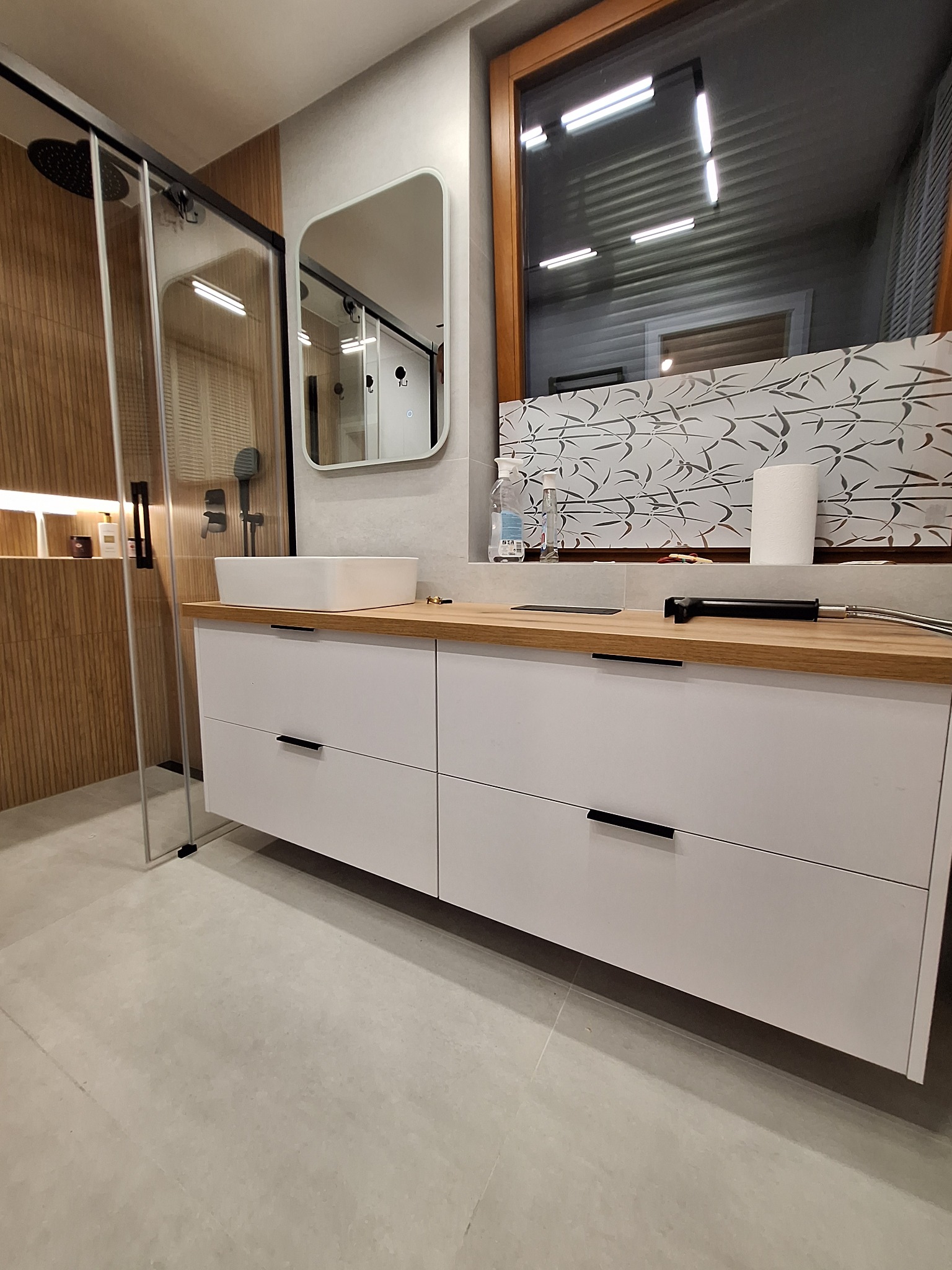 Nowoczesna łazienka z białą szafką pod umywalką z drewnianym blatem, kabiną prysznicową i lustrem. Minimalistyczny design, jasne kolory, bambusowy motyw na ścianie.