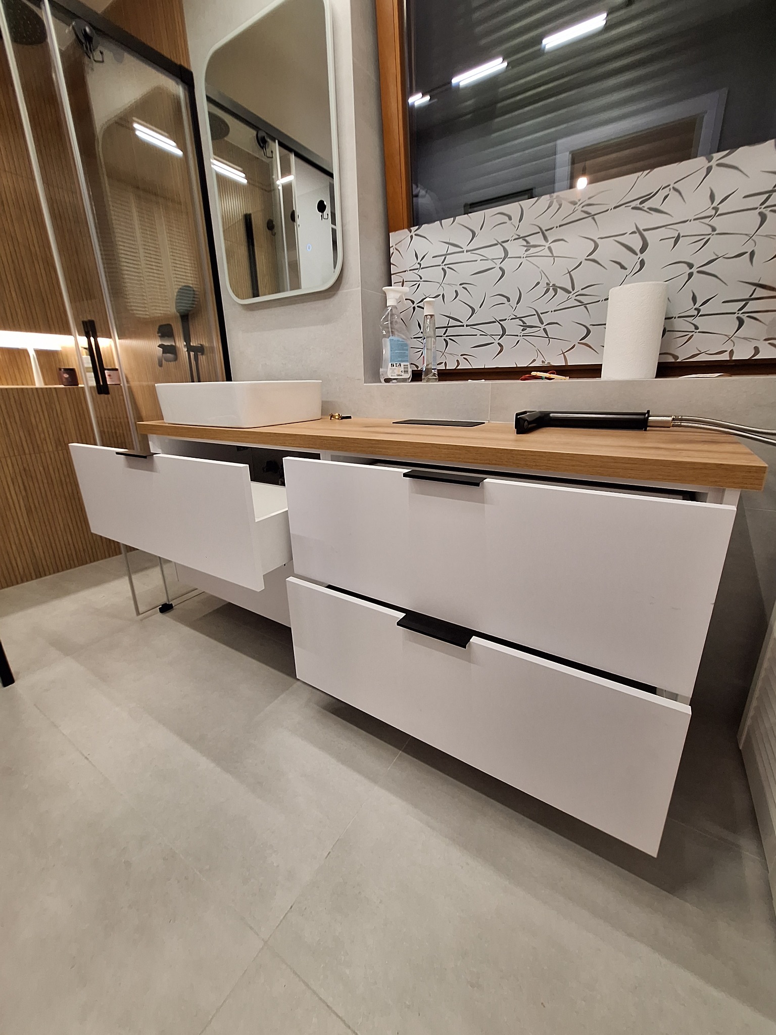 Nowoczesna łazienka z białą szafką pod umywalkę z drewnianym blatem i czarnymi uchwytami. Widoczna kabina prysznicowa i lustro. Otwarta szuflada.