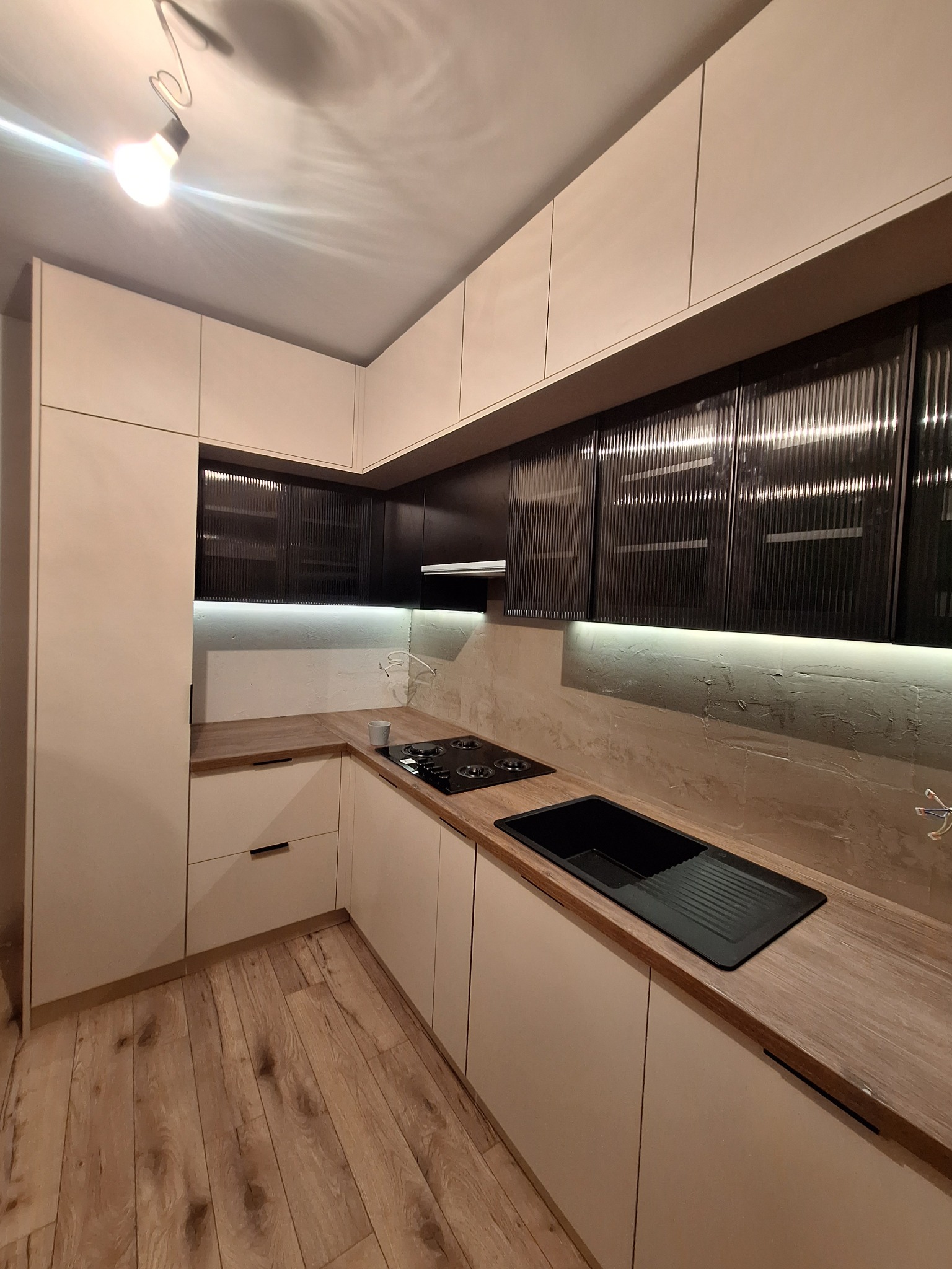 Nowoczesna kuchnia z beżowymi szafkami i czarnymi ryflowanymi frontami górnymi. Blat drewnopodobny, oświetlenie LED podszafkowe. Minimalistyczny design.