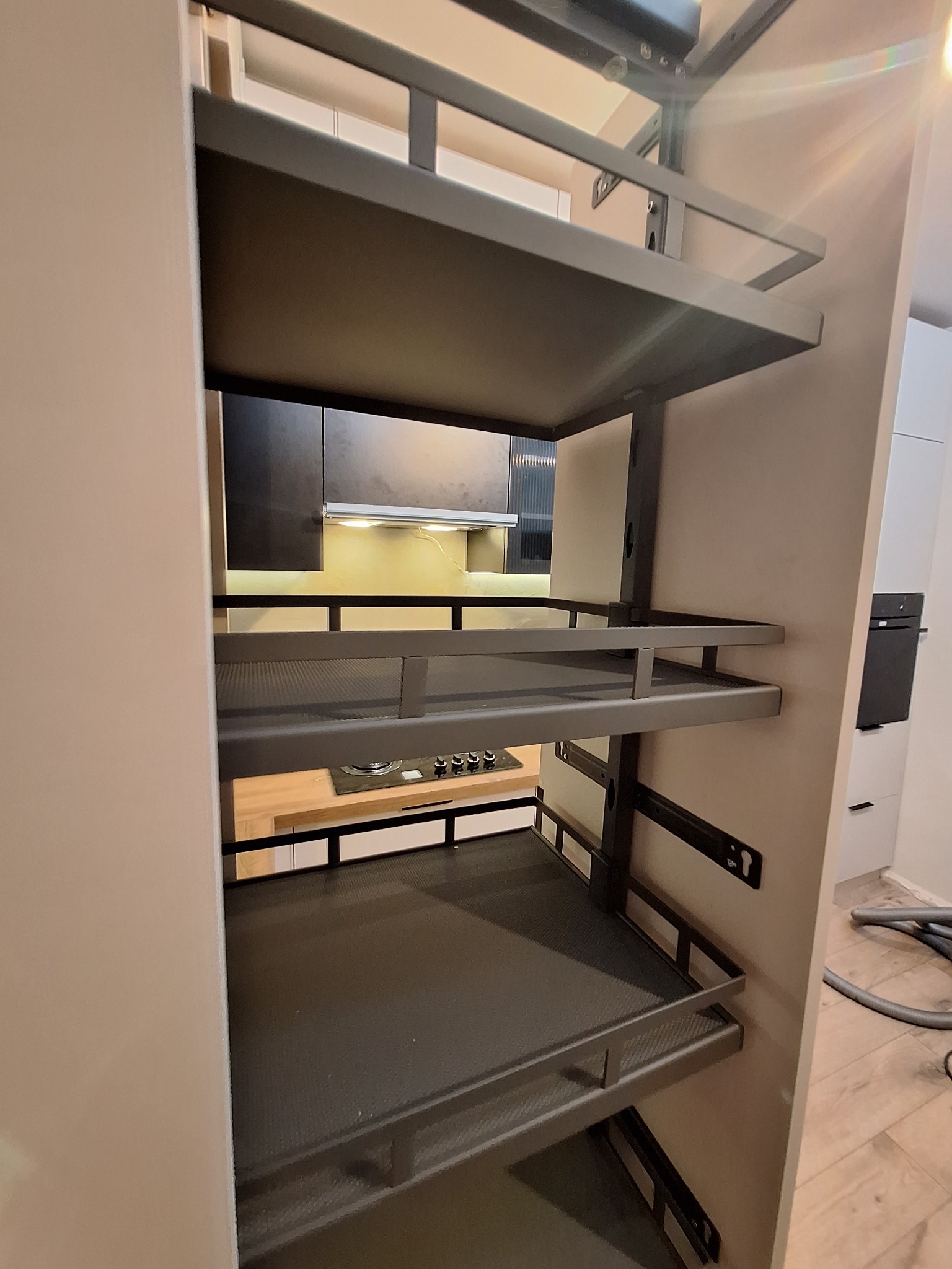 Wysuwana, metalowa półka cargo w szafce kuchennej, widok na płytę grzewczą i czarne szafki w tle. Nowoczesne rozwiązanie do przechowywania w kuchni.