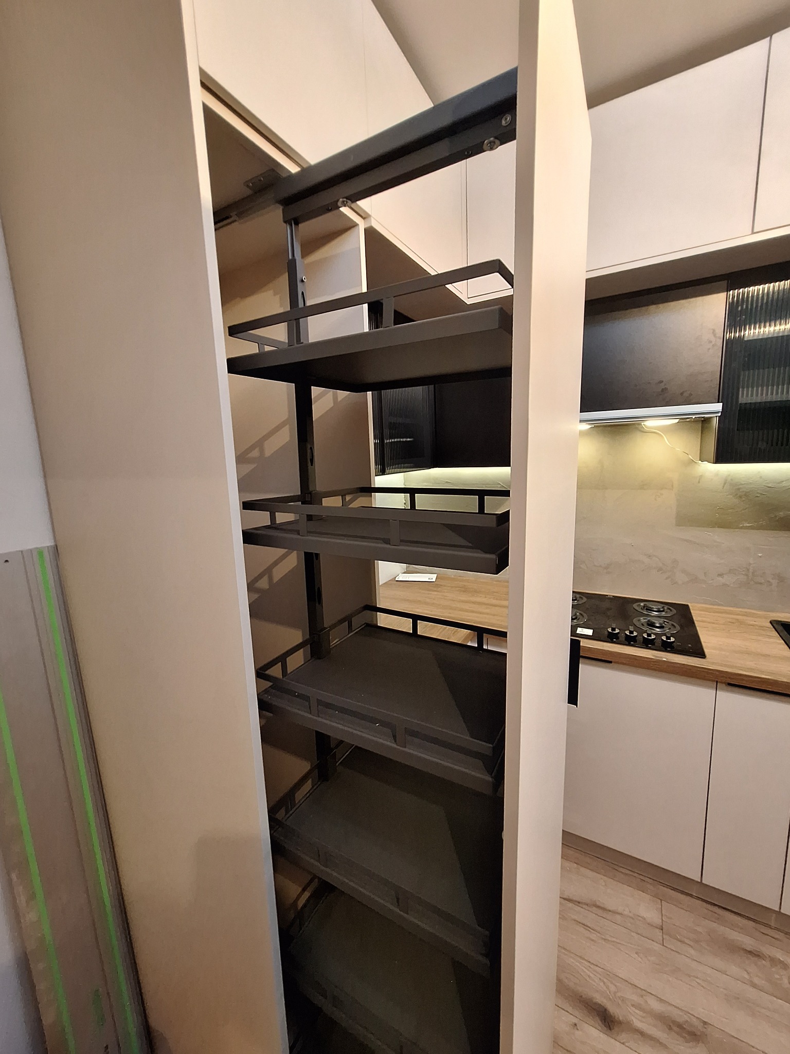 Wysuwana, metalowa półka cargo w zabudowie kuchennej na wymiar. Widoczne półki z relingami, jasne fronty szafek i blat z płyty drewnopodobnej.